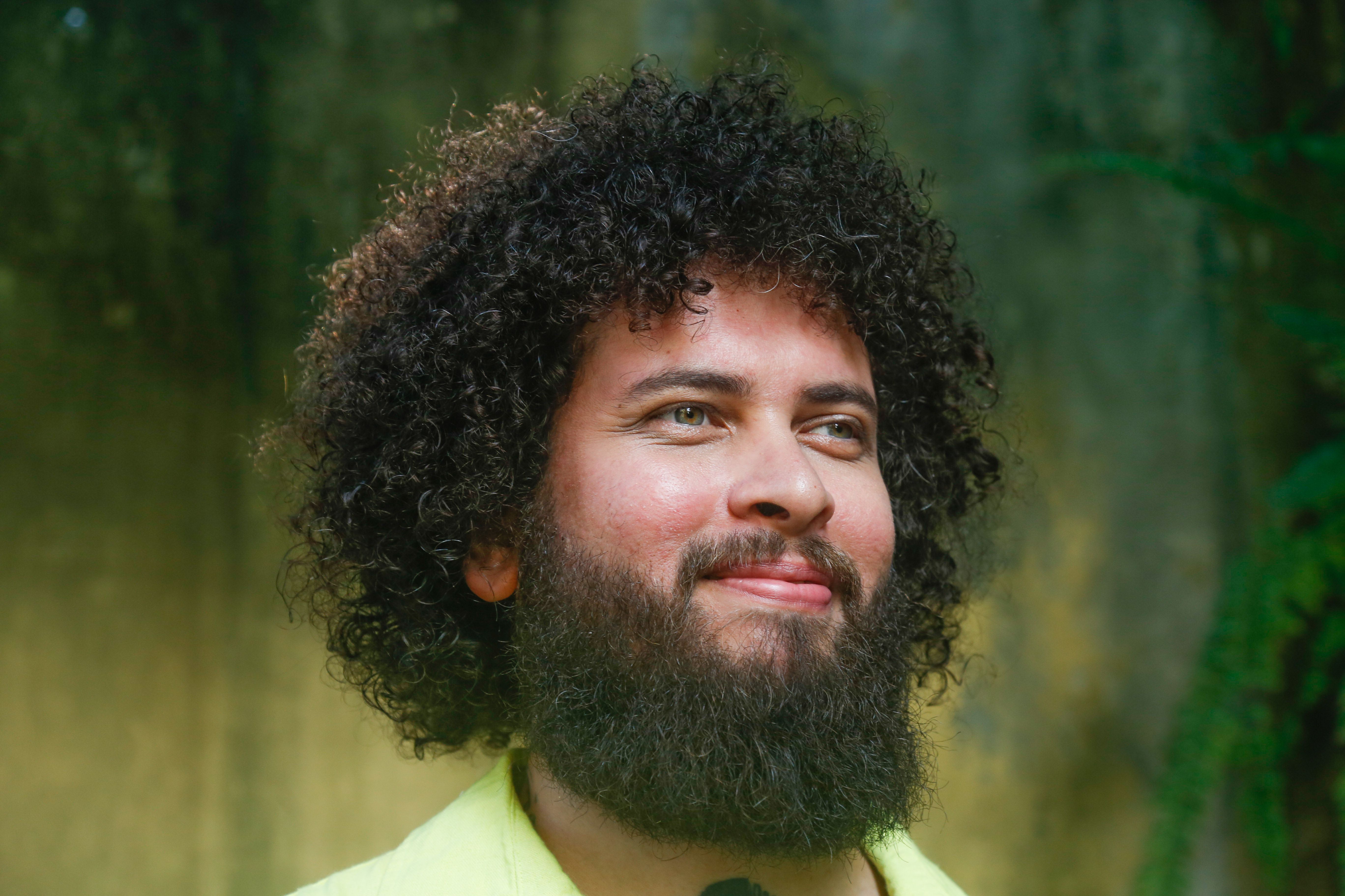 Retrato em close-up de um homem de pele clara com um volumoso cabelo crespo em estilo black power e barba cheia e escura. Ele tem olhos claros e um leve sorriso, olhando para o lado e para cima com uma express&atilde;o serena. Veste uma pe&ccedil;a de roupa em tom verde-lim&atilde;o claro. O fundo est&aacute; desfocado, sugerindo um ambiente externo com vegeta&ccedil;&atilde;o e tons esverdeados e terrosos. A luz &eacute; suave, destacando os detalhes de sua express&atilde;o e a textura dos fios de cabelo