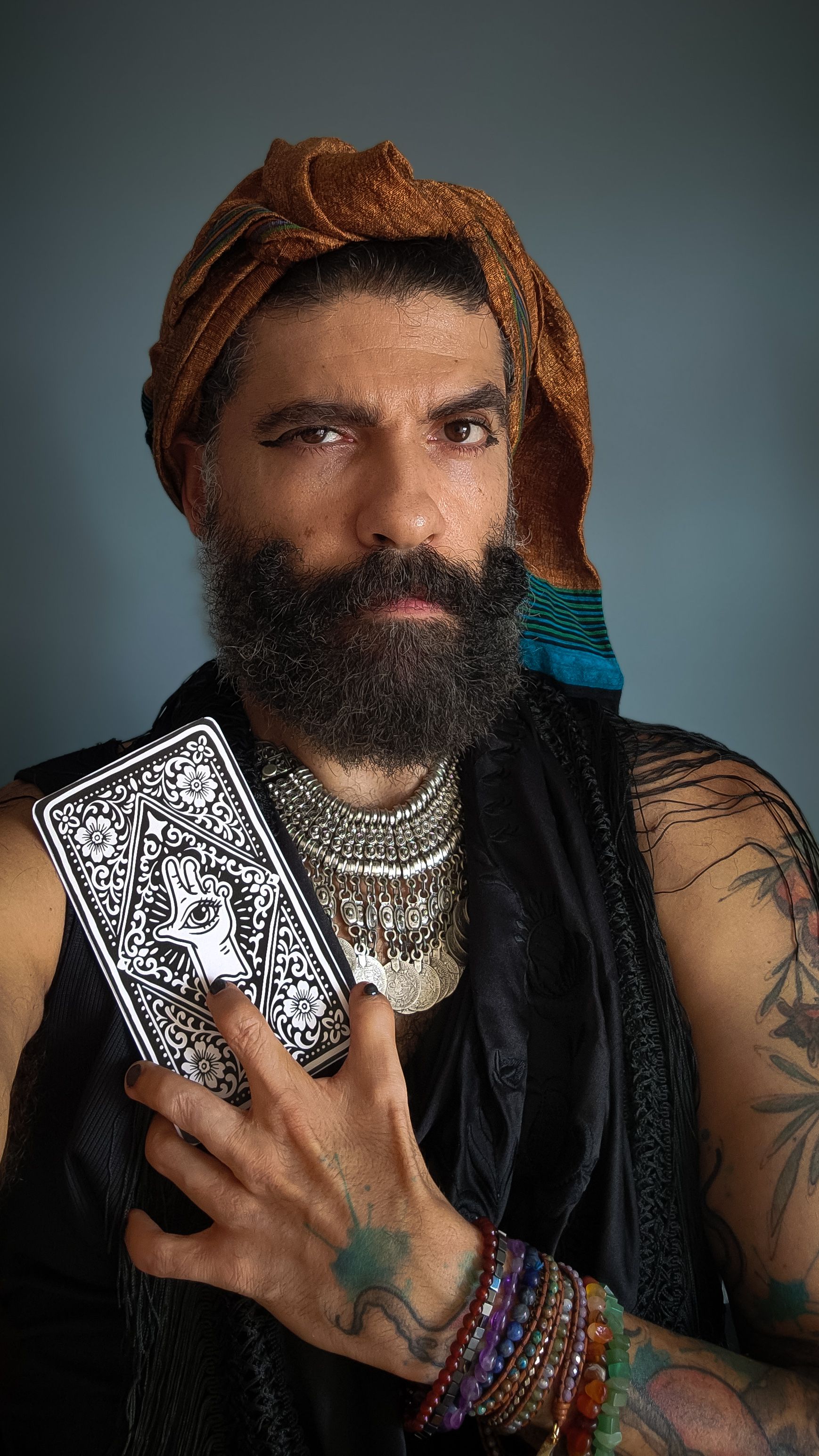 Um autorretrato vertical em plano m&eacute;dio mostra um homem de pele clara e barba longa e escura. Ele veste um turbante ocre, roupas pretas e diversos colares prateados de estilo &eacute;tnico. Seus bra&ccedil;os est&atilde;o cobertos por tatuagens coloridas e o pulso exibe v&aacute;rias pulseiras de pedras. Com a m&atilde;o esquerda, ele segura contra o peito uma carta de tar&ocirc; com ilustra&ccedil;&otilde;es em preto e branco que destacam uma m&atilde;o com um olho. Seus olhos est&atilde;o real&ccedil;ados por maquiagem preta, e ele encara a c&acirc;mera com express&atilde;o enigm&aacute;tica. O fundo &eacute; de um tom cinza-azulado s&oacute;lido
