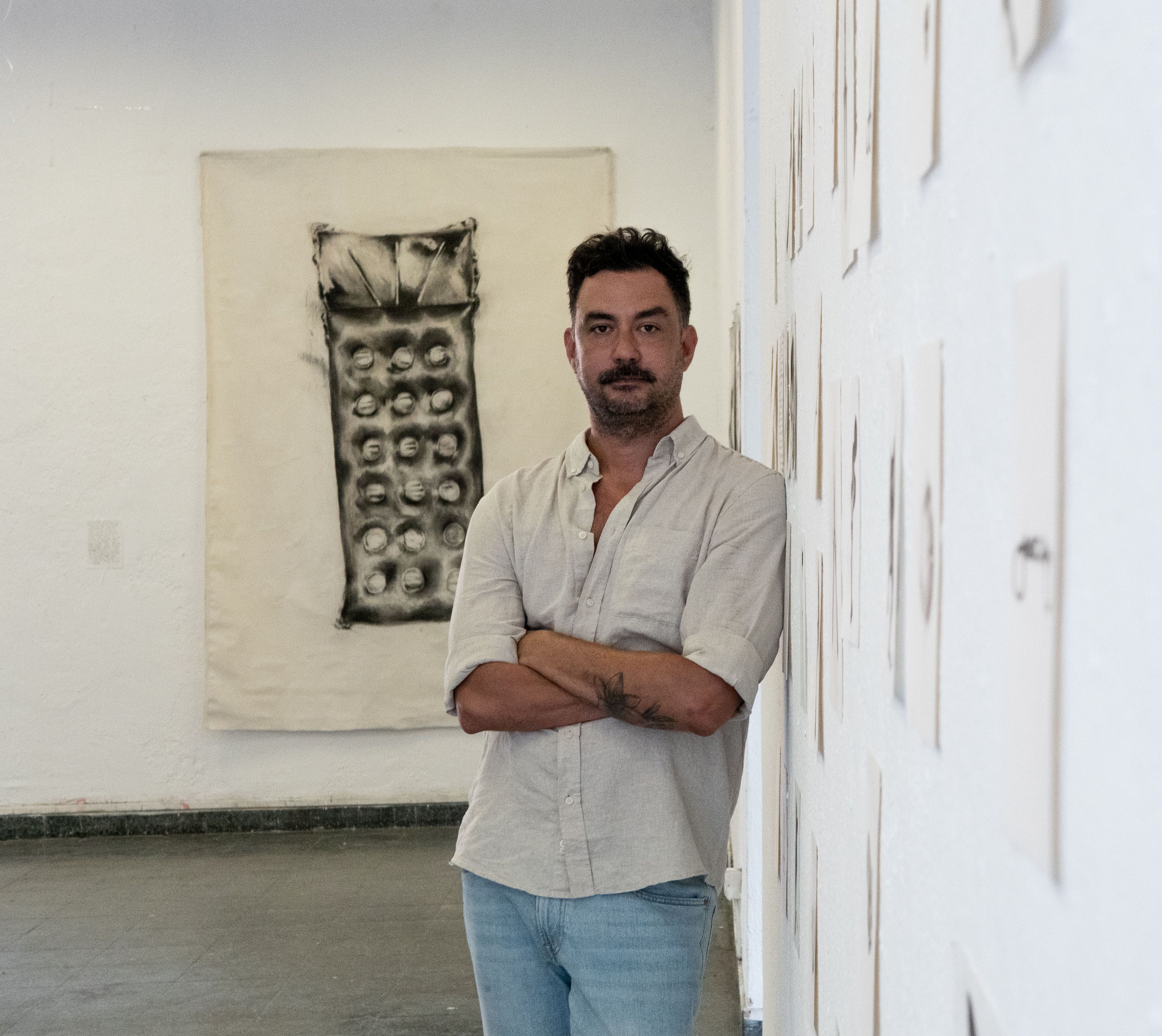 Uma fotografia em plano m&eacute;dio, com fundo em tons de cinza e branco, mostram um homem de pele clara e cabelos curtos e escuros. Ele veste uma camisa cinza de mangas longas e cal&ccedil;a azul-escura e est&aacute; apoiado em uma parede branca com os bra&ccedil;os cruzados, encarando a c&acirc;mera com seriedade. Ao fundo, uma grande pe&ccedil;a de tape&ccedil;aria branca est&aacute; pendurada na parede, com um desenho em preto de uma cama e travesseiros. &Agrave; direita, pequenos desenhos brancos e pretos est&atilde;o pendurados na parede, e a luz natural entra pela janela lateral