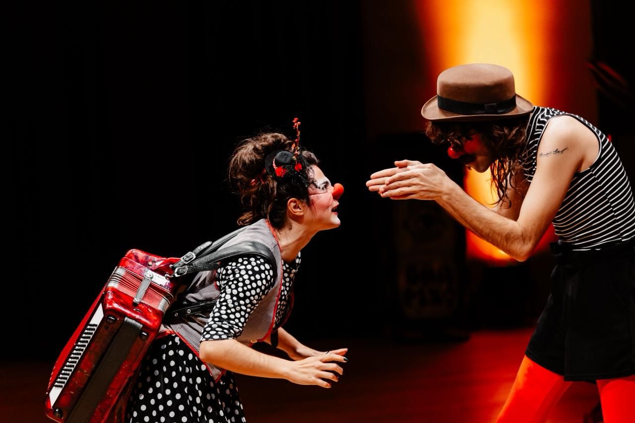 
Duas figuras de palha&ccedil;aria interagem em um palco escuro com luz quente ao fundo. &Agrave; esquerda, uma mulher de pele clara, nariz de palha&ccedil;o vermelho e maquiagem art&iacute;stica encara o parceiro. Ela usa uma roupa de bolinhas pretas e brancas e carrega um acorde&atilde;o vermelho nas costas. &Agrave; direita, um homem de chap&eacute;u coco marrom, nariz de palha&ccedil;o e camiseta listrada inclina-se para frente, com as m&atilde;os estendidas em dire&ccedil;&atilde;o a ela. Ele veste cal&ccedil;as curtas pretas sobre uma meia-cal&ccedil;a vermelha vibrante. A cena &eacute; din&acirc;mica e expressiva
