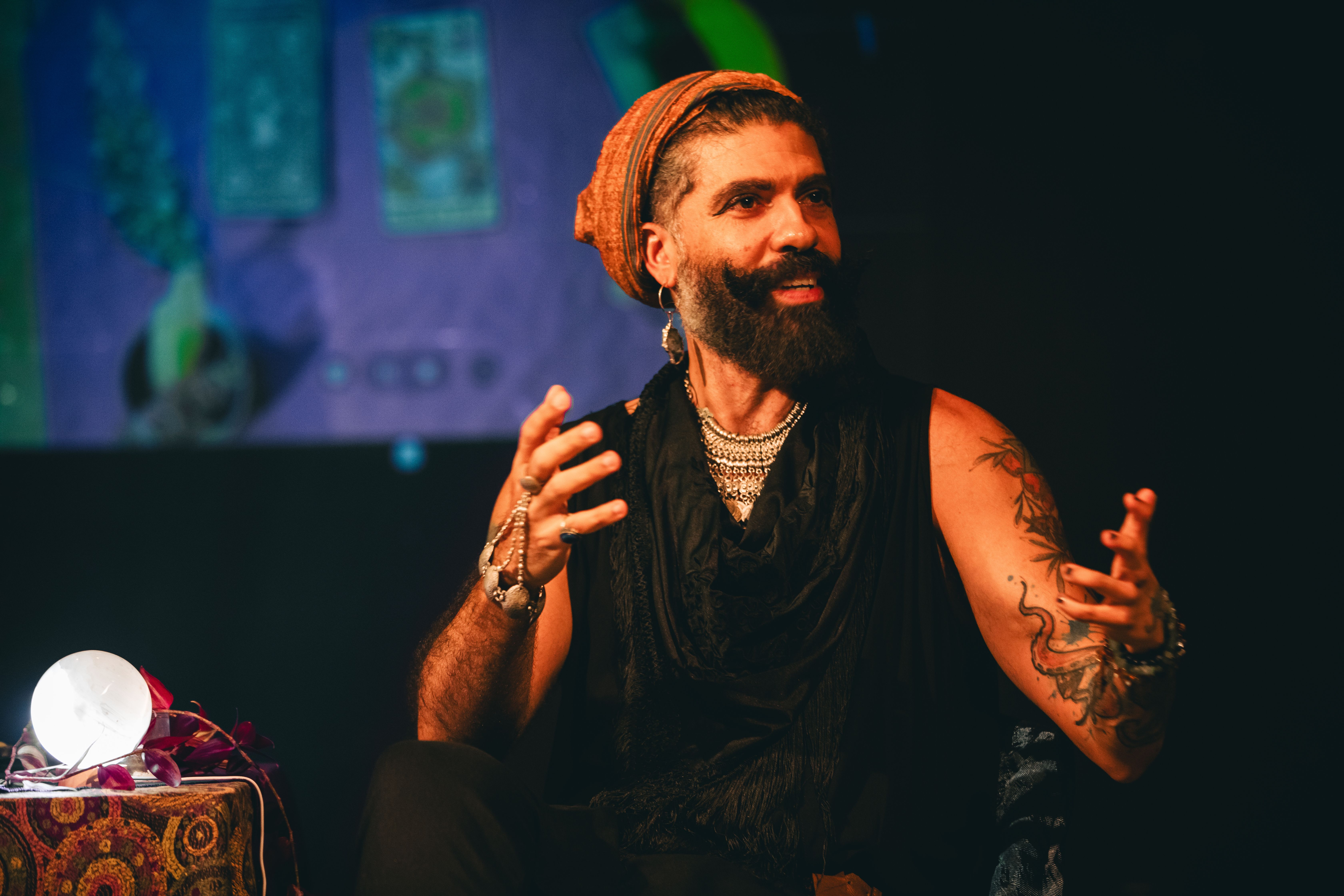 Fotografia em plano m&eacute;dio de um homem durante uma performance. Ele tem pele clara com barba longa e escura, vestindo um turbante ocre e roupas pretas. Seus bra&ccedil;os est&atilde;o tatuados e ele usa diversos colares prateados e acess&oacute;rios nas m&atilde;os. Ele est&aacute; sentado, gesticulando com as m&atilde;os abertas enquanto olha para o lado com um leve sorriso. &Agrave; esquerda, em primeiro plano, h&aacute; uma mesa com uma bola de cristal iluminada e flores roxas. Ao fundo, uma proje&ccedil;&atilde;o desfocada mostra cartas de tar&ocirc;. A ilumina&ccedil;&atilde;o &eacute; quente e teatral