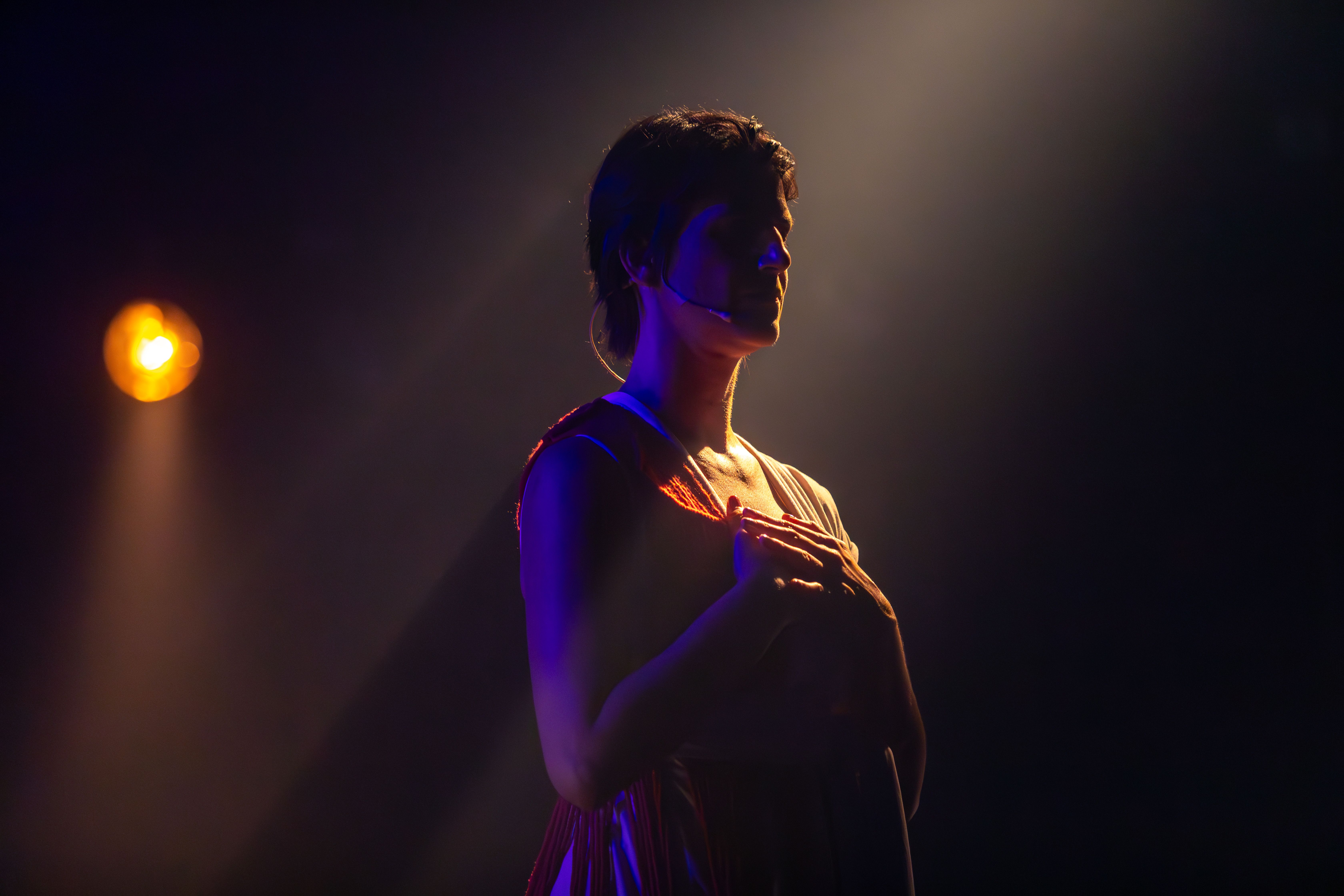 Fotografia de close-up lateral de uma mulher de pele clara e cabelos curtos em um palco escuro. Ela est&aacute; com os olhos fechados e as m&atilde;os cruzadas sobre o peito, em uma express&atilde;o de introspec&ccedil;&atilde;o. A ilumina&ccedil;&atilde;o &eacute; dram&aacute;tica: um feixe de luz &acirc;mbar incide por tr&aacute;s, contornando sua silhueta com um brilho dourado, enquanto uma luz azulada ilumina suavemente sua nuca e o microfone de cabe&ccedil;a. &Agrave; esquerda, um refletor circular amarelado aparece desfocado ao fundo. O contraste entre luz e sombra acentua o clima solene e po&eacute;tico da cena