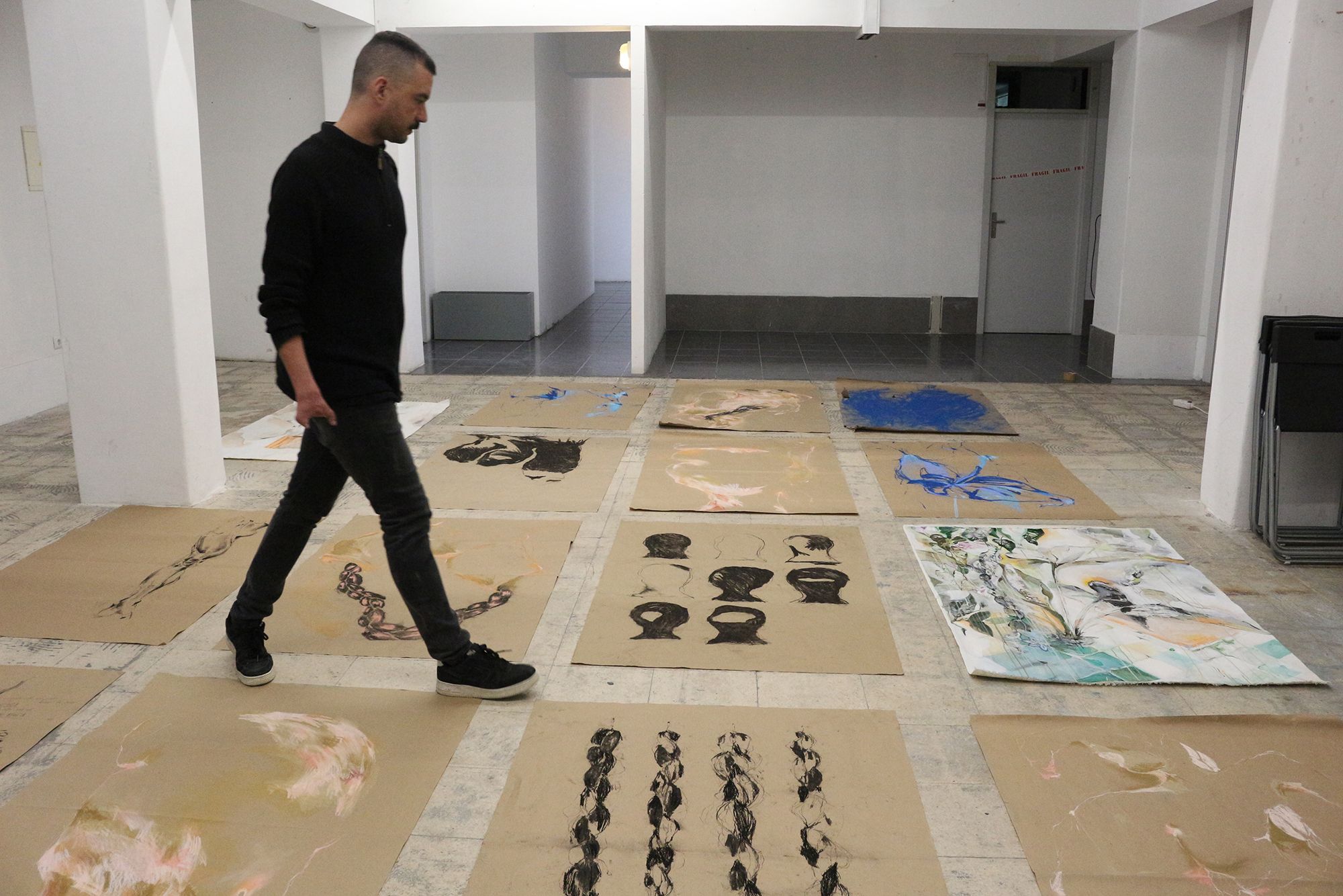 Um homem de pele clara, vestindo blusa preta e cal&ccedil;a jeans, caminha da esquerda para a direita em uma galeria de arte. Ele tem cabelos curtos, raspados nas laterais. O ch&atilde;o do espa&ccedil;o amplo e branco est&aacute; forrado com diversas folhas grandes de papel pardo e telas, contendo desenhos manuais com formas, rostos e linhas, predominantemente em tons de preto, azul e rosado. O homem atravessa o espa&ccedil;o entre as obras dispostas pelo ch&atilde;o. O ambiente &eacute; iluminado, com paredes brancas.