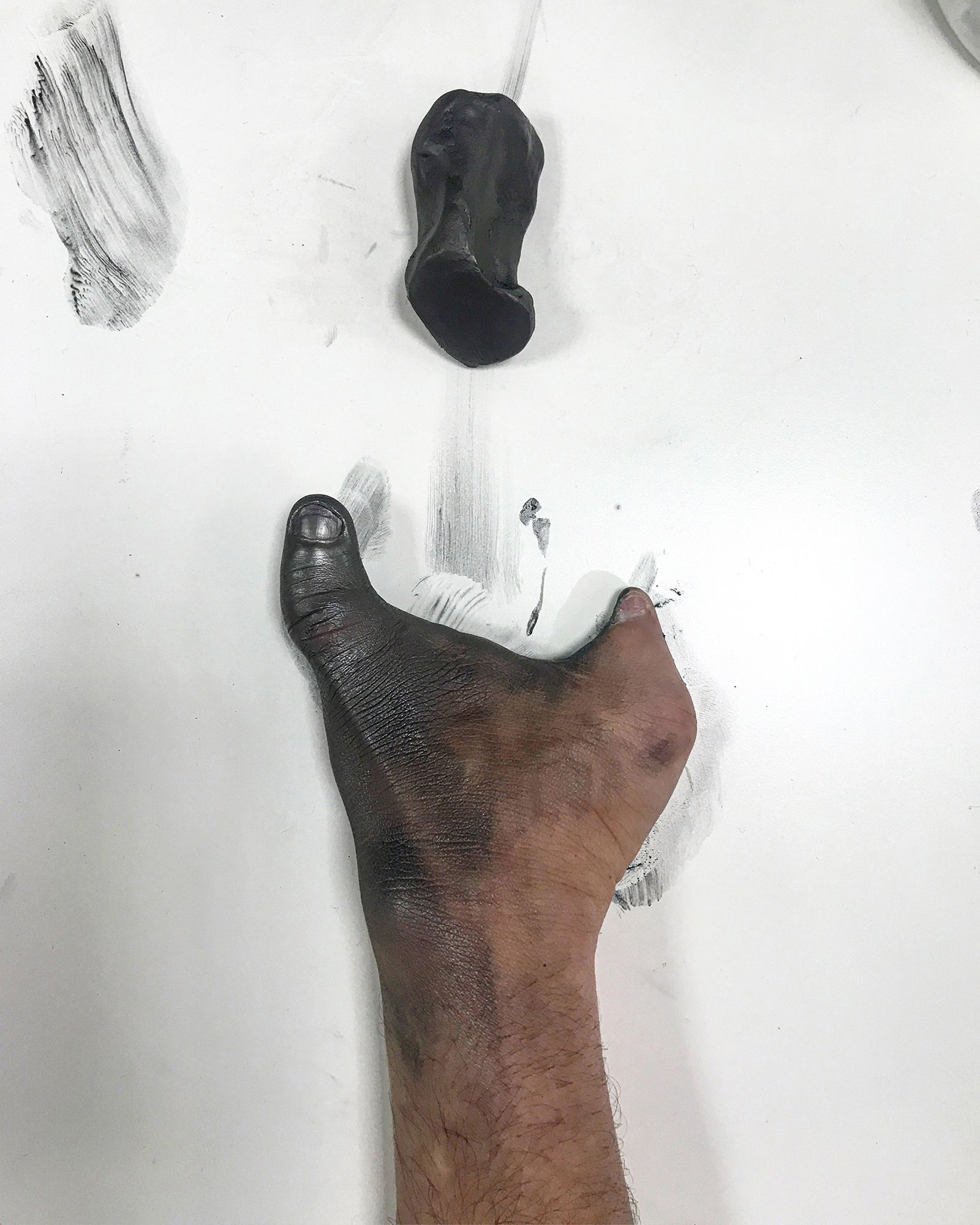 Foto em close-up de um bra&ccedil;o e m&atilde;o sobre uma superf&iacute;cie branca. A m&atilde;o &eacute; parcialmente amputada e a pele est&aacute; coberta por uma subst&acirc;ncia preta e densa, semelhante a carv&atilde;o ou pigmento, que se concentra do punho at&eacute; o polegar. A m&atilde;o est&aacute; aberta e estendida. Acima da m&atilde;o, na superf&iacute;cie branca, h&aacute; um pequeno mont&iacute;culo da mesma subst&acirc;ncia preta, com formato irregular e denso. O fundo &eacute; uma superf&iacute;cie branca com marcas de rastro da mesma subst&acirc;ncia, sugerindo um processo de manuseio ou cria&ccedil;&atilde;o art&iacute;stica manual.