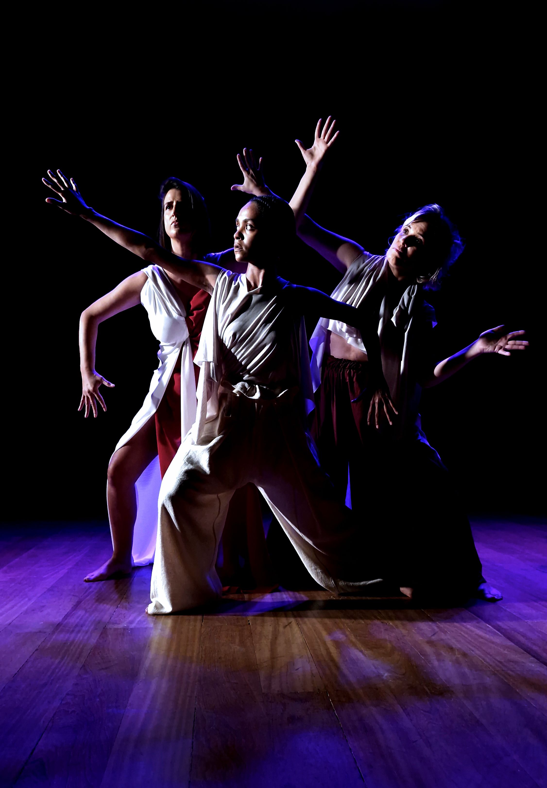 Tr&ecirc;s mulheres em um palco de madeira, capturadas em uma pose expressiva de dan&ccedil;a. Elas vestem roupas em tons de branco e vinho, com tecidos fluidos que acompanham seus movimentos. A dan&ccedil;arina ao centro est&aacute; &agrave; frente, com os joelhos dobrados e bra&ccedil;os abertos, enquanto as outras duas, nas laterais, estendem os bra&ccedil;os em dire&ccedil;&otilde;es opostas. O fundo &eacute; completamente escuro e a ilumina&ccedil;&atilde;o &eacute; dram&aacute;tica, em tons de azul e roxo, destacando as formas e express&otilde;es das int&eacute;rpretes. A cena transmite for&ccedil;a, dramaticidade e movimento art&iacute;stico.