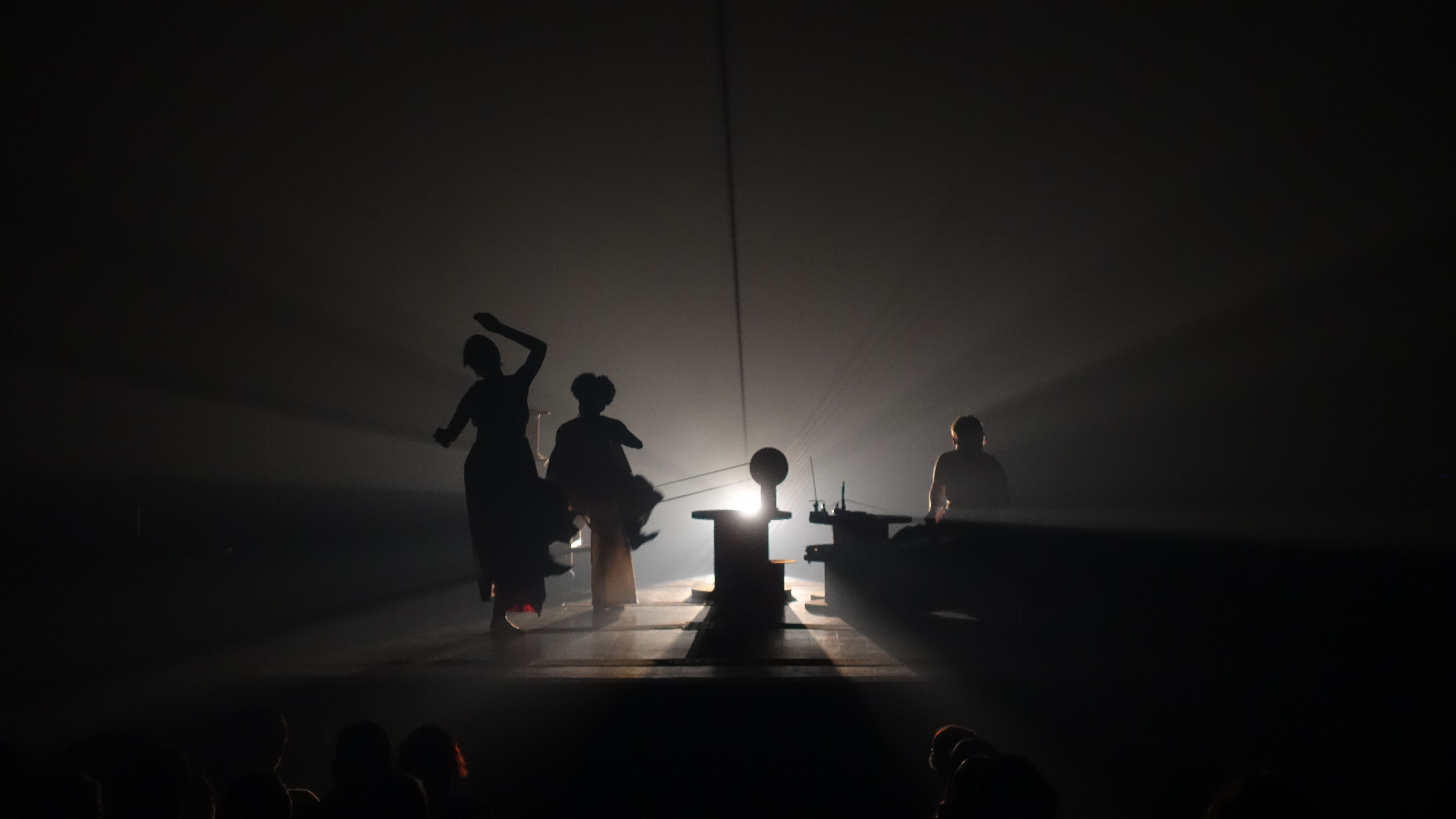 Vista ampla de um palco de teatro em penumbra. Tr&ecirc;s silhuetas humanas aparecem em destaque contra uma luz intensa que emana do centro do palco, criando feixes de luz que cortam o ambiente. No centro, h&aacute; um pedestal ou instrumento. As figuras est&atilde;o em movimento, sugerindo uma performance. Na parte inferior da imagem, v&ecirc;-se a silhueta das cabe&ccedil;as da plateia, indicando que o ponto de vista &eacute; do p&uacute;blico. A atmosfera &eacute; misteriosa, dram&aacute;tica e teatral, com forte contraste entre a luz intensa de fundo e as sombras das figuras.