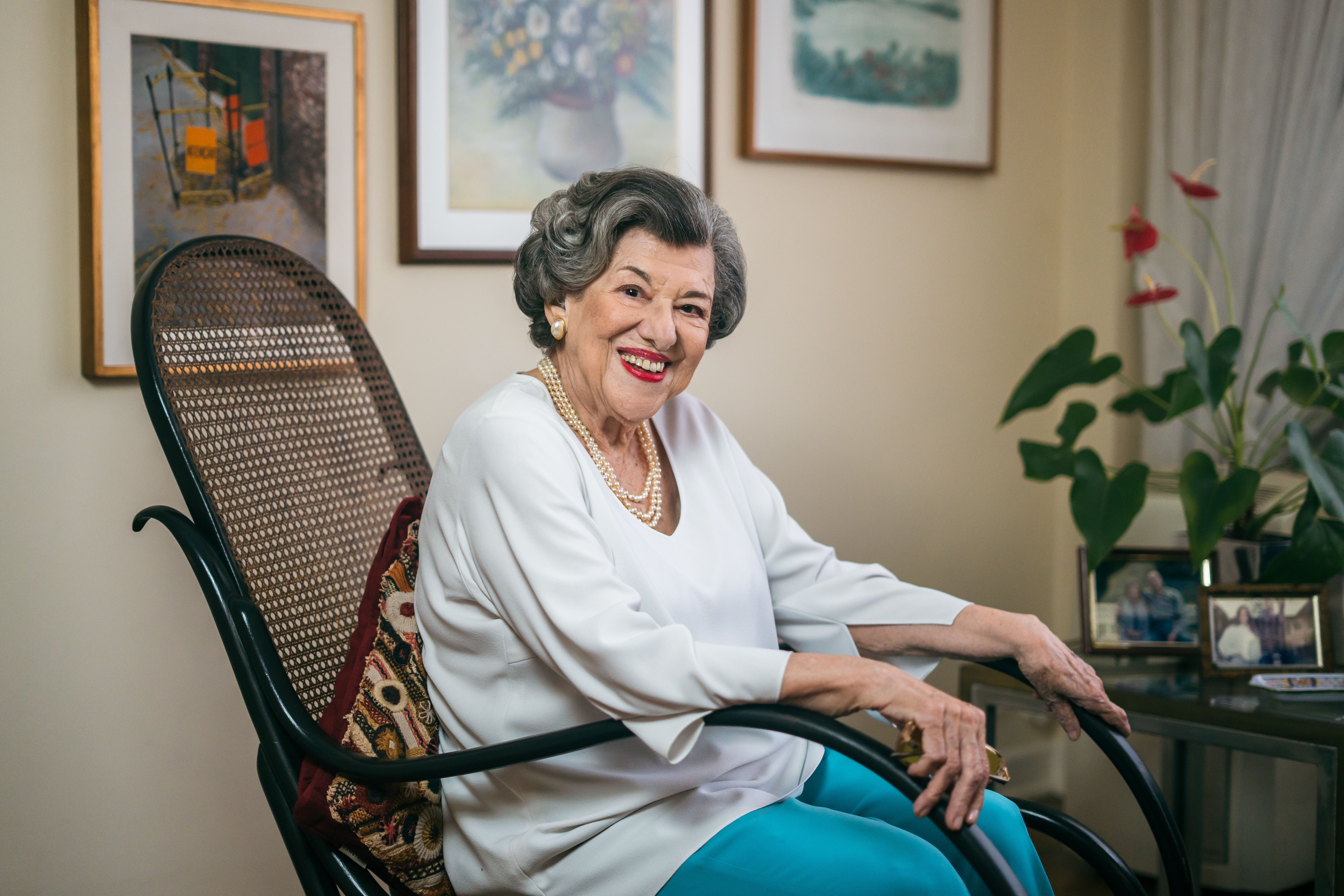 Ruth Rocha, senhora branca de cabelos grisalhos e sorriso aberto, senta-se em uma cadeira de balanço de palhinha preta. Veste blusa branca, colar de pérolas e calça azul-turquesa. Ao fundo, quadros decoram a parede clara e há um vaso com antúrios vermelhos à direita.