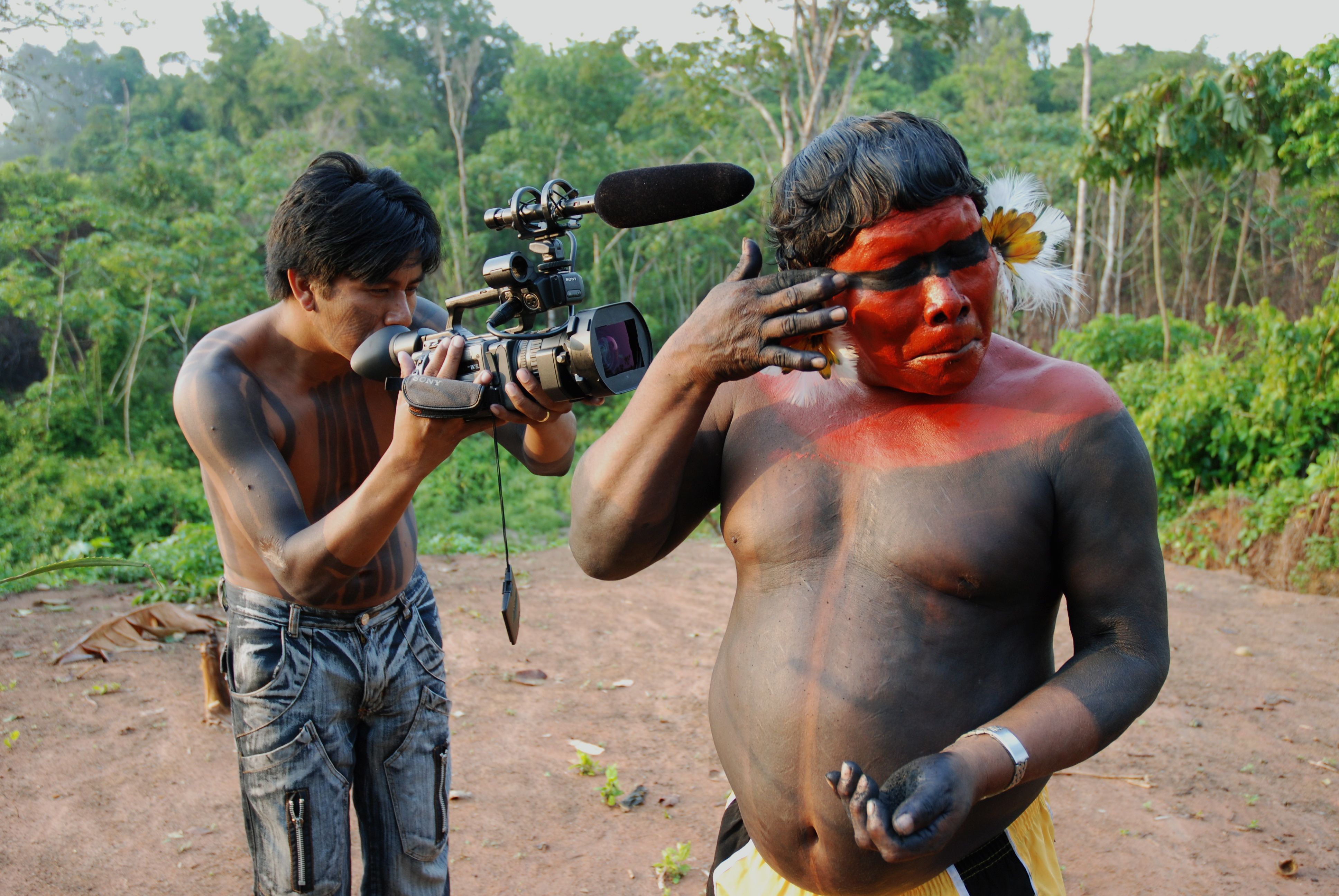 Há dois homens indígenas em meio a um cenário de mata. Ambos estão sem camiseta e possuem pinturas típicas em seu tronco. Um segura uma câmera e filma o outro, que está com o rosto vermelho e, no momento da foto, está passando uma tinta preta sobre os olhos - que estão fechados - de forma horizontal. 