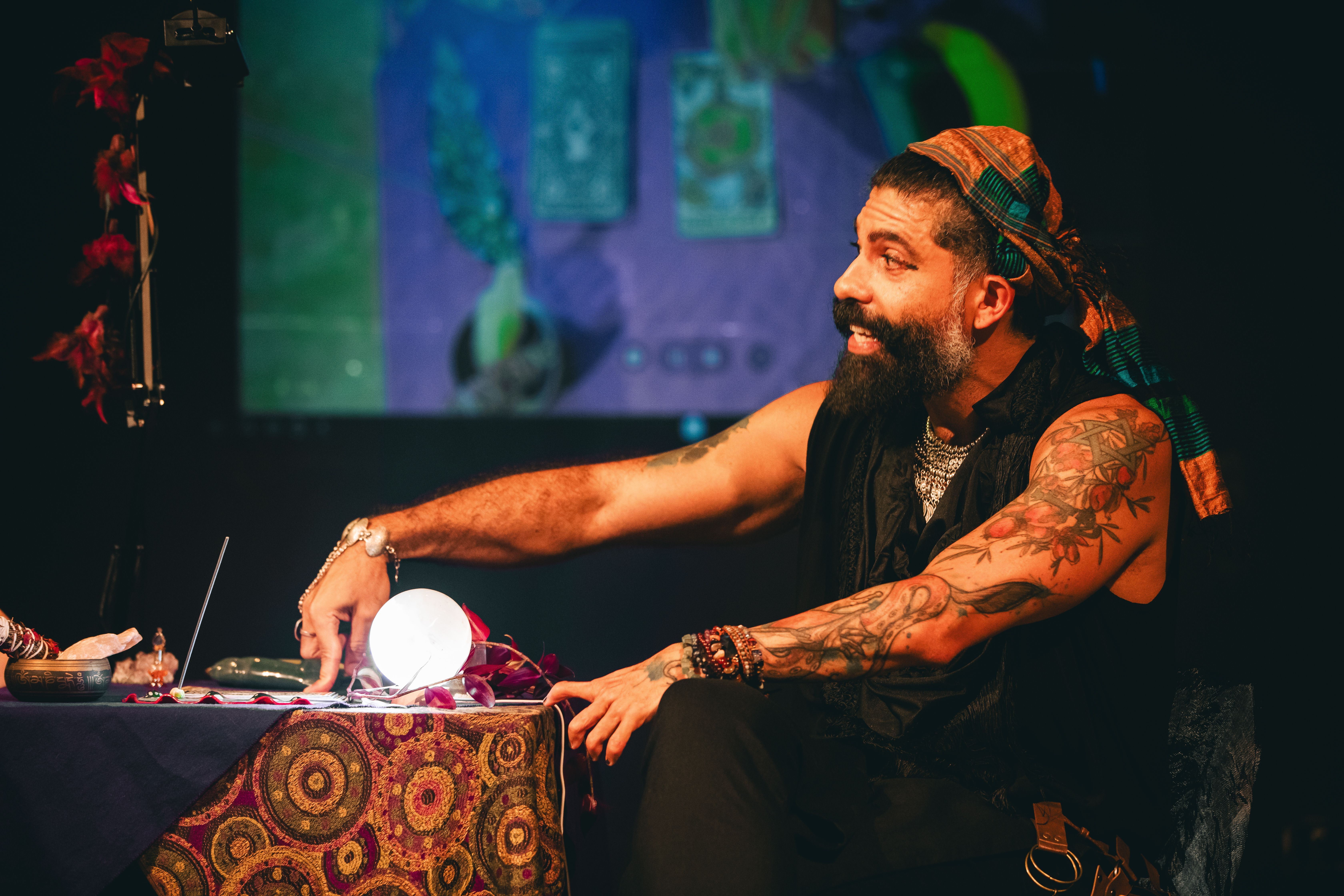 Homem de pele parda e barba escura, sentado &agrave; frente de uma mesa de tar&ocirc;. Ele veste preto, usa um len&ccedil;o colorido na cabe&ccedil;a e possui diversas tatuagens nos bra&ccedil;os. Sua express&atilde;o &eacute; animada, com olhos arregalados e um leve sorriso. Sobre a mesa com toalha estampada, h&aacute; uma esfera de luz branca intensa, cristais e uma tigela tibetana. Ao fundo, uma proje&ccedil;&atilde;o desfocada exibe cartas de tar&ocirc; em tons de azul e verde. A ilumina&ccedil;&atilde;o &eacute; dram&aacute;tica, destacando a figura do homem contra o cen&aacute;rio escuro. No canto esquerdo, h&aacute; flores vermelhas presas a um suporte.