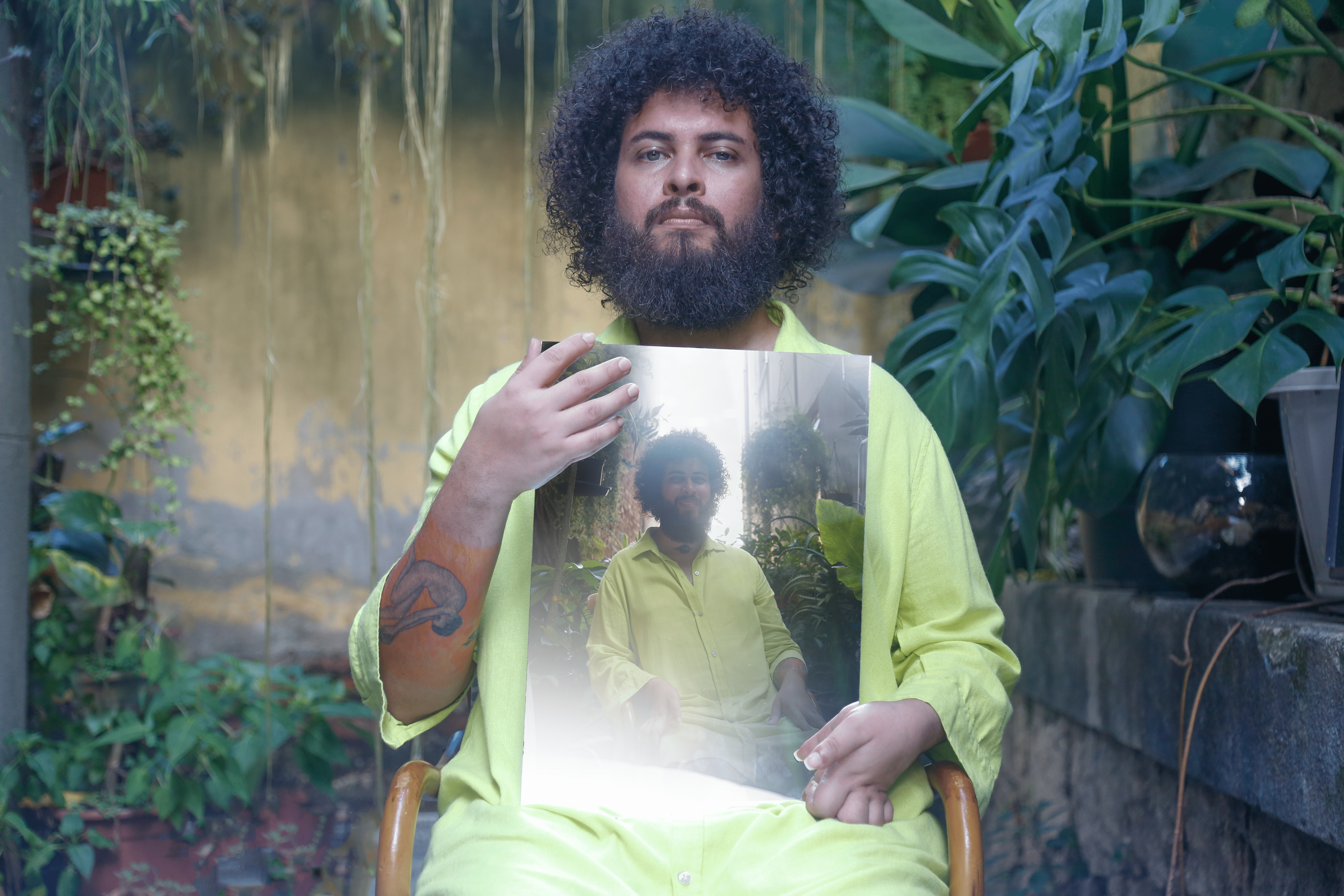 Um homem de cabelos crespos e camisa verde-lim&atilde;o est&aacute; sentado em um jardim com diversas plantas ao fundo, incluindo uma costela-de-ad&atilde;o. Ele segura, na altura do peito, uma fotografia impressa de si mesmo na mesma pose e ambiente. Em seu bra&ccedil;o direito, uma tatuagem colorida &eacute; vis&iacute;vel. O cen&aacute;rio externo tem tons de verde e paredes r&uacute;sticas, criando uma composi&ccedil;&atilde;o de 