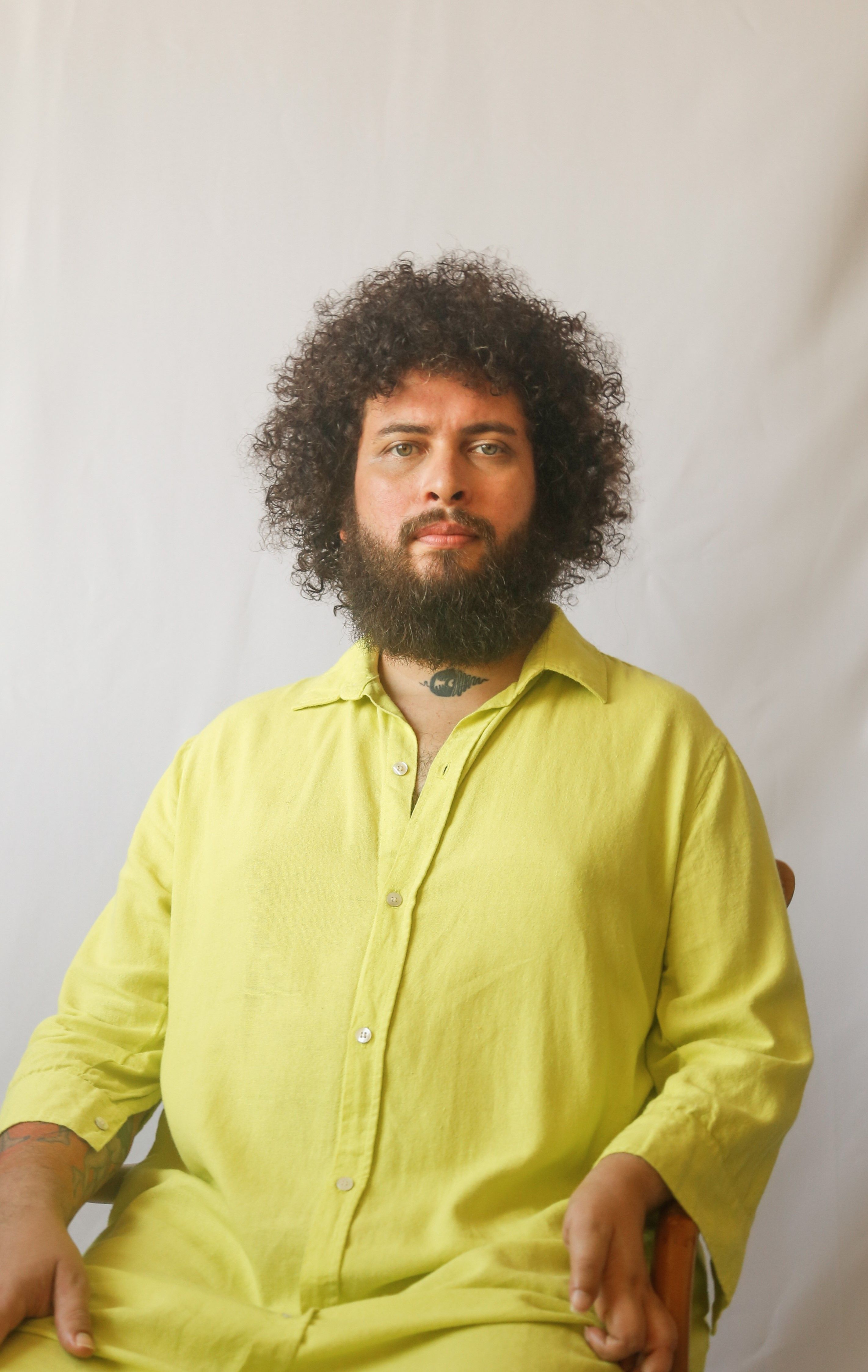 Retrato de um homem de pele parda, com cabelos crespos volumosos e barba densa e escura. Ele olha diretamente para a c&acirc;mera com uma express&atilde;o serena. Veste uma camisa de linho em tom verde-lim&atilde;o vibrante. No centro do pesco&ccedil;o, possui uma pequena tatuagem escura. O fundo &eacute; branco e minimalista, destacando a textura do tecido da roupa e os detalhes do rosto. A ilumina&ccedil;&atilde;o &eacute; suave e frontal.