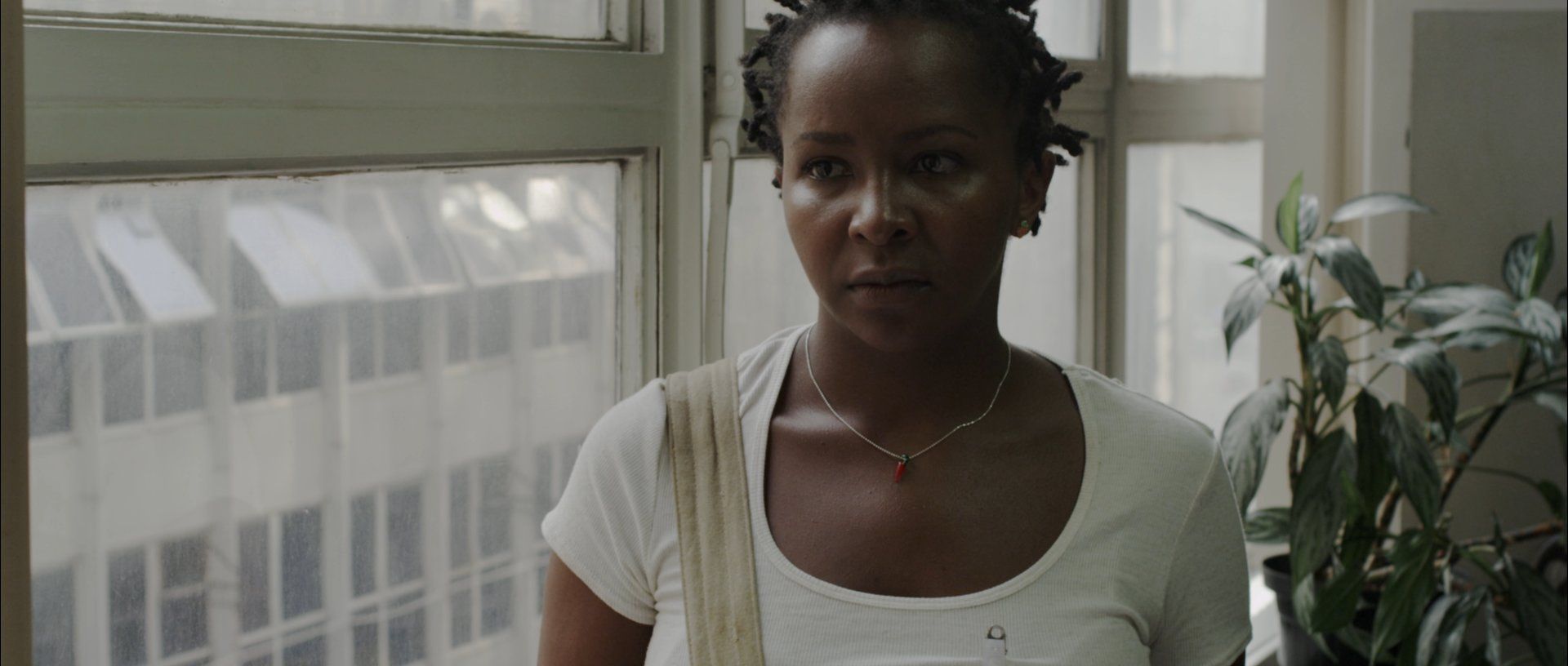 A imagem mostra a atriz Débora Marçal em cena. Ela é uma mulher negra e veste uma blusa branca. Atrás dela, há janelas de vidro e uma planta.