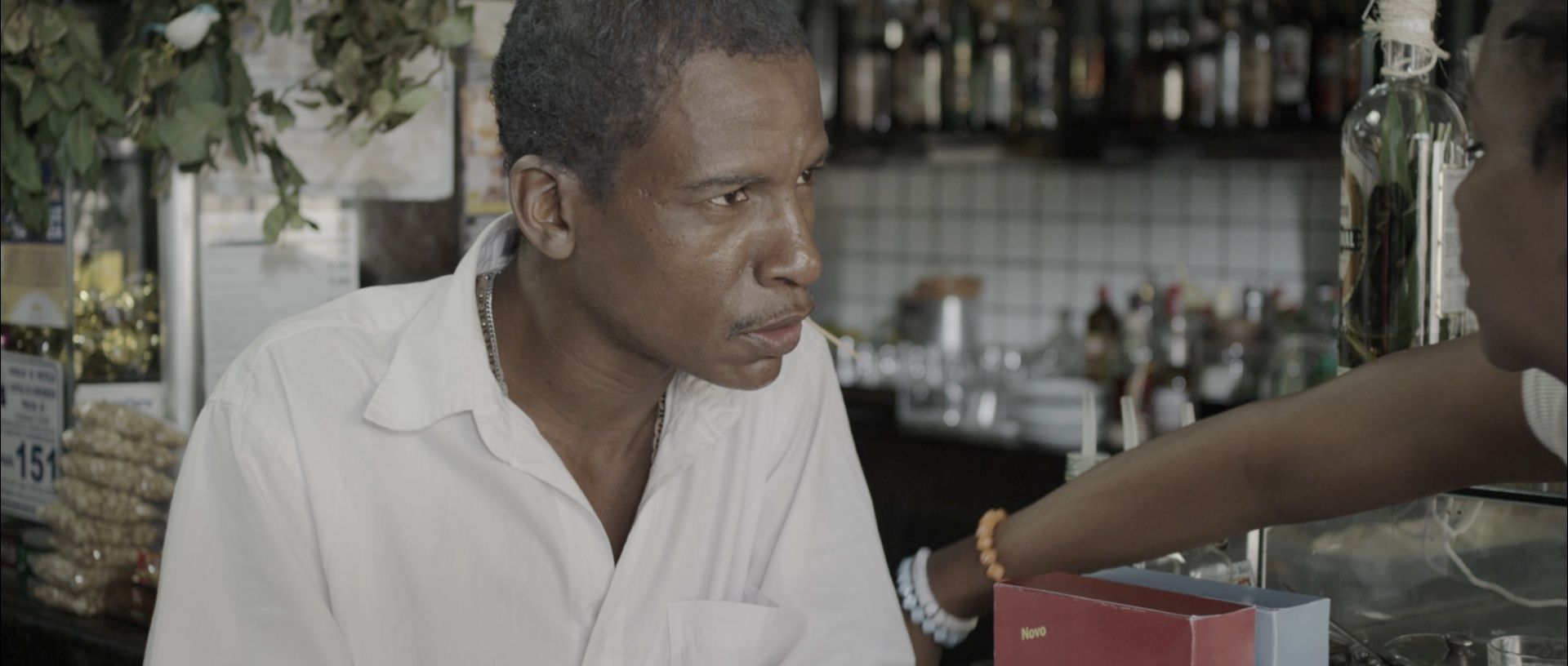 Um homem negro, usando uma camisa branca, est&aacute; em um bar. Ele olha em dire&ccedil;&atilde;o a uma atendente.