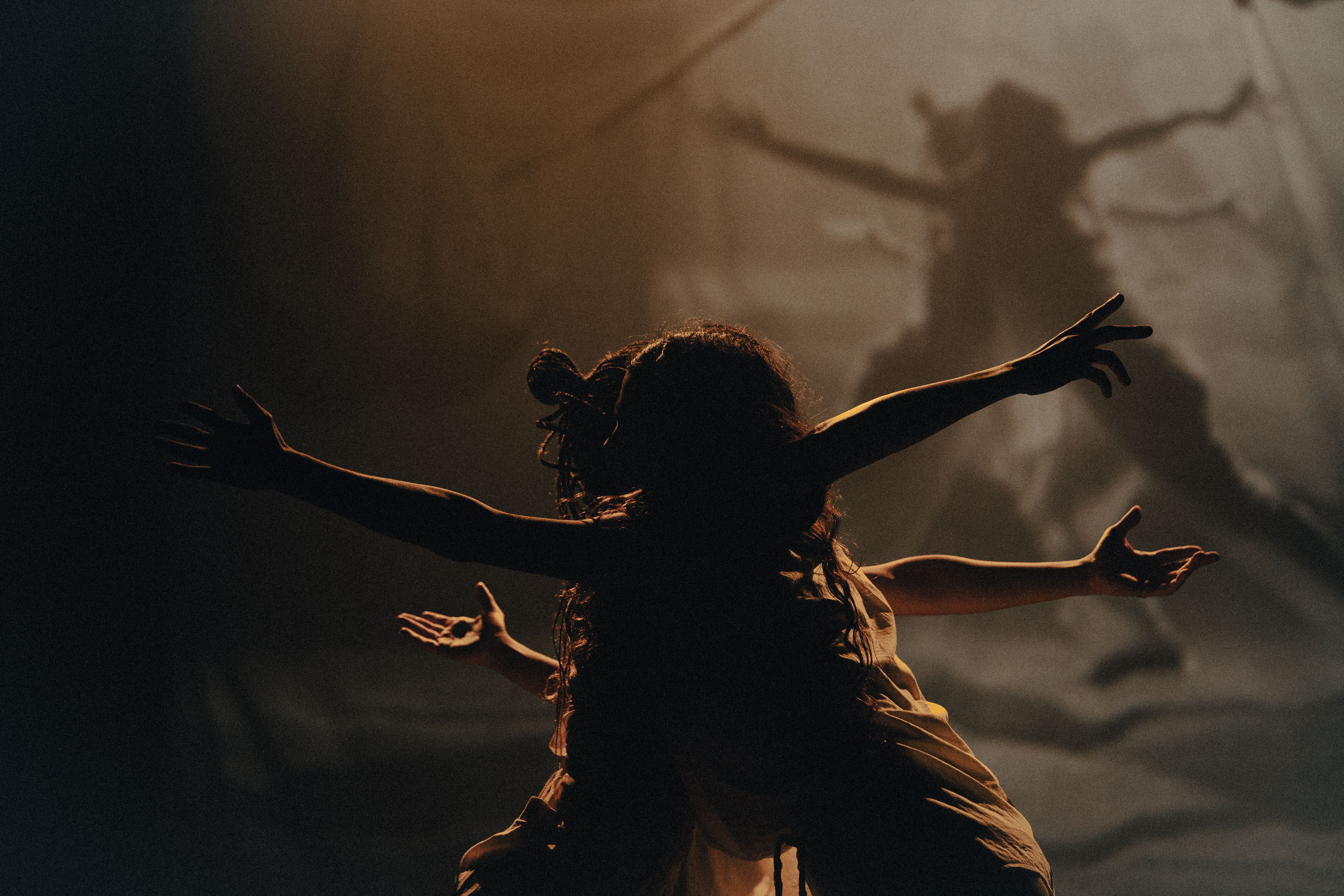 Fotografia em plano m&eacute;dio com ilumina&ccedil;&atilde;o dram&aacute;tica em tons de s&eacute;pia e &acirc;mbar. Duas pessoas se abra&ccedil;am e mant&eacute;m os bra&ccedil;os esticados na horizontal, criando o efeito visual de uma figura com m&uacute;ltiplos bra&ccedil;os, como uma divindade. Ao fundo, uma proje&ccedil;&atilde;o difusa mostra uma sombra gigante que mimetiza a pose, amplificando a sensa&ccedil;&atilde;o de expans&atilde;o e movimento ancestral no palco.