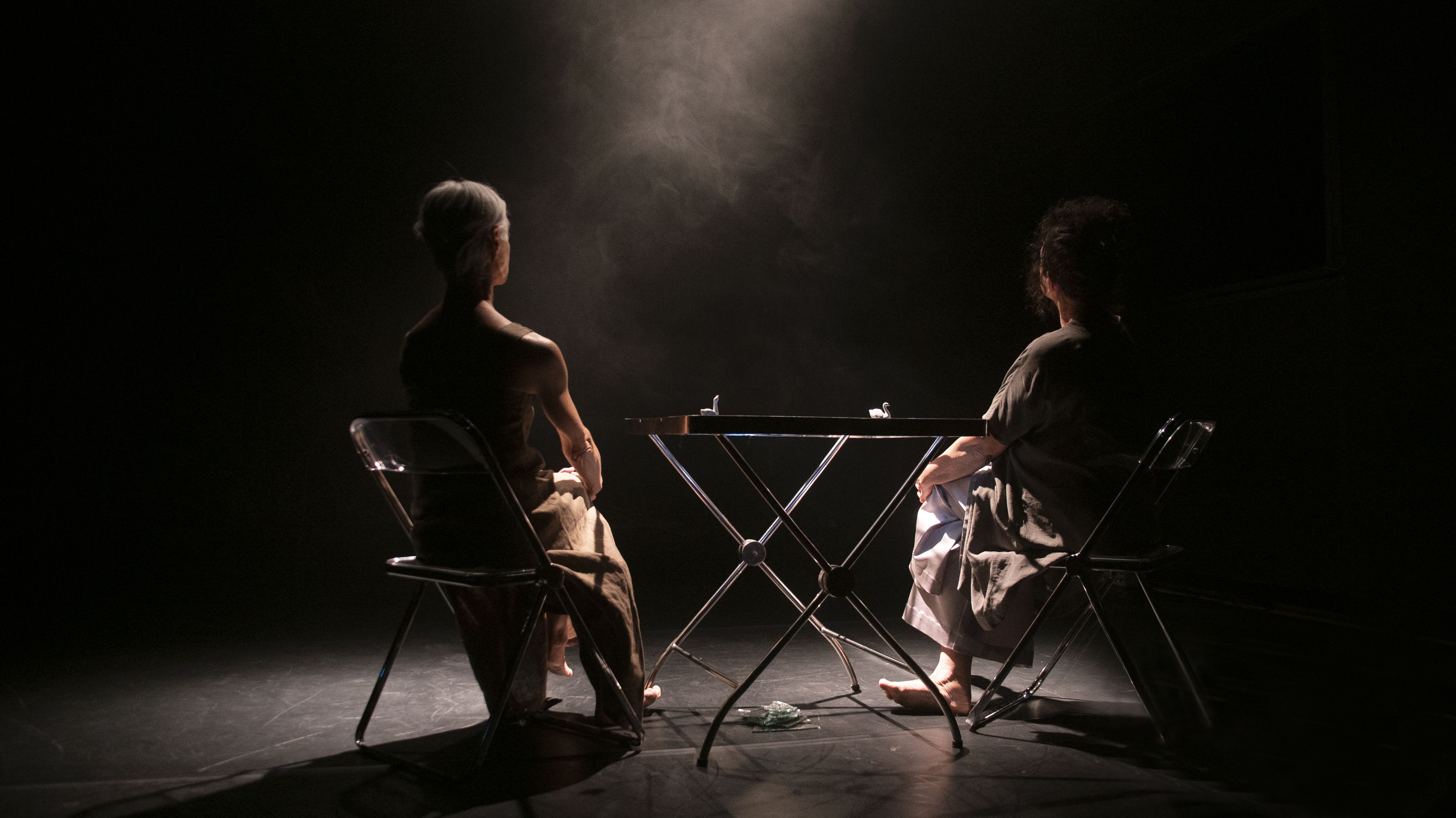 Fotografia horizontal de um palco &agrave;s escuras. No centro, duas mulheres est&atilde;o sentadas em cadeiras de metal dobr&aacute;veis, de costas para a c&acirc;mera, separadas por uma pequena mesa dobr&aacute;vel. Elas s&atilde;o iluminadas por um &uacute;nico foco de luz vertical que incide sobre a mesa, revelando uma leve fuma&ccedil;a no ar. A mulher &agrave; esquerda tem cabelos curtos e grisalhos; a da direita tem cabelos escuros e volumosos. O clima &eacute; de introspec&ccedil;&atilde;o, sil&ecirc;ncio e um encontro &iacute;ntimo em meio ao vazio.