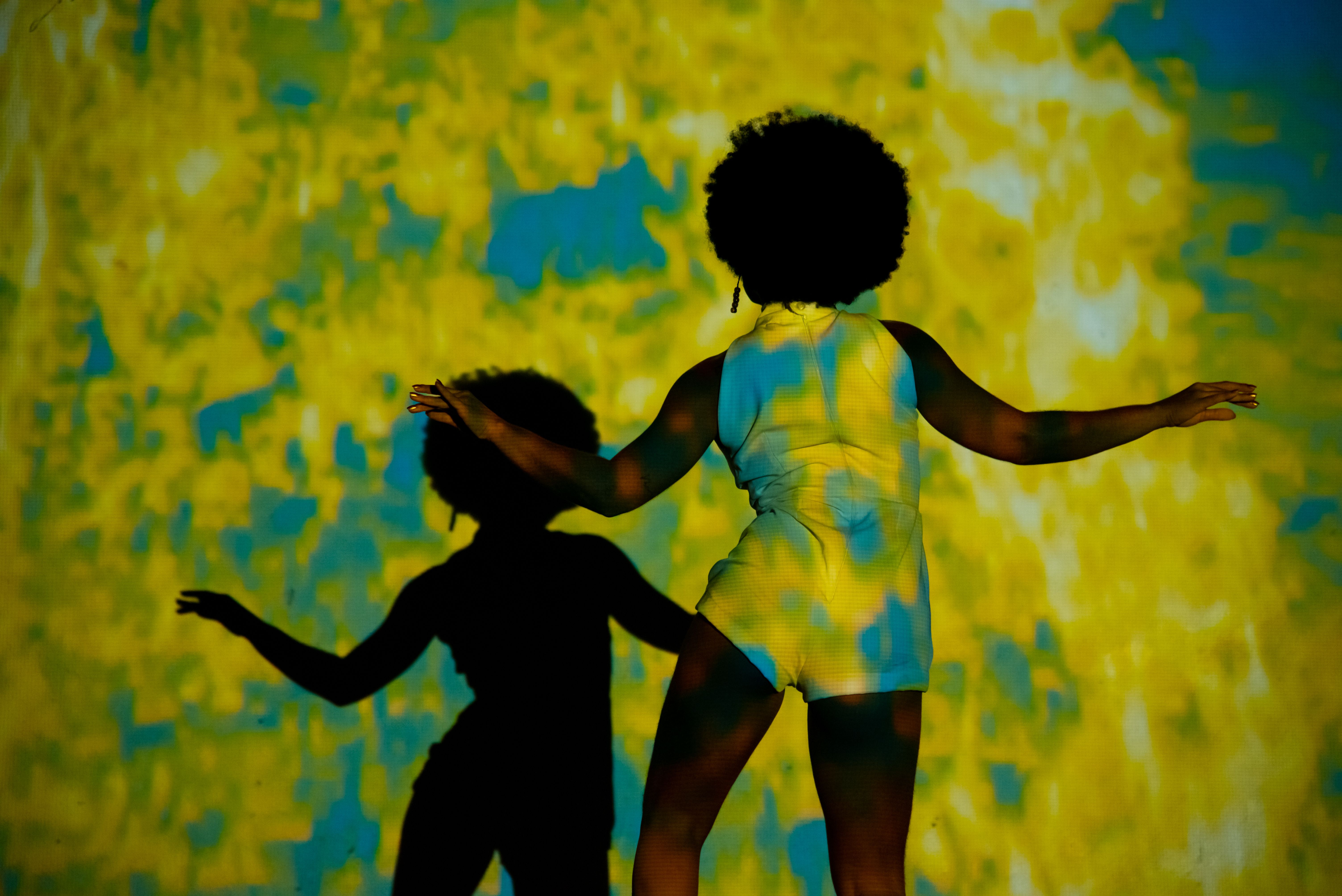 Fotografia vibrante e colorida. Uma mulher negra de costas, com cabelo estilo black power, ocupa o centro da imagem. Ela veste um macac&atilde;o curto claro. &Agrave; sua frente, uma parede recebe a proje&ccedil;&atilde;o de manchas abstratas em azul e amarelo, que tamb&eacute;m cobrem seu corpo. &Agrave; esquerda, sua sombra &eacute; projetada de forma n&iacute;tida, criando um contraste direto entre o corpo f&iacute;sico iluminado e a silhueta escura. Seus bra&ccedil;os est&atilde;o levemente flexionados para os lados, em posi&ccedil;&atilde;o de dan&ccedil;a.
