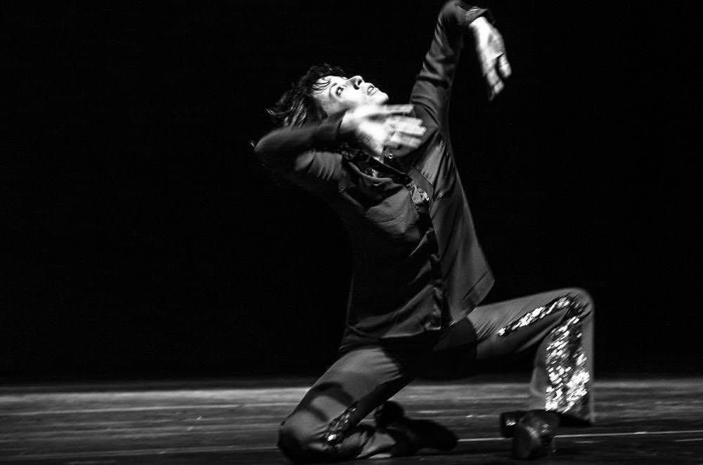 Fotografia em preto e branco de alta expressividade. Uma pessoa bailarina est&aacute; ajoelhada no palco, com o corpo inclinado para tr&aacute;s em uma pose din&acirc;mica. Veste uma camisa escura e cal&ccedil;as com detalhes brilhantes nas laterais. O bra&ccedil;o direito est&aacute; erguido acima da cabe&ccedil;a com o punho flexionado, enquanto o esquerdo est&aacute; pr&oacute;ximo ao rosto em movimento, gerando um leve rastro de desfoque. A express&atilde;o facial &eacute; intensa, voltada para cima, contra um fundo totalmente escuro.