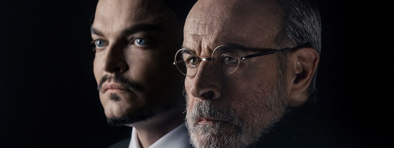 Em um fundo preto e ilumina&ccedil;&atilde;o dram&aacute;tica de penumbra, o ator Osmar Prado aparece em primeiro plano. Ele &eacute; um homem idoso, branco, de barba grisalha, usa &oacute;culos redondos e veste um traje escuro de gola alta. Logo atr&aacute;s dele, parcialmente vis&iacute;vel, est&aacute; o ator Maur&iacute;cio Machado, um homem branco, jovem, com olhos azuis marcantes, cabelo escuro penteado e bigode fino, vestindo terno e gravata. Ambos olham s&eacute;rios e concentrados para a esquerda, fora da c&acirc;mera.