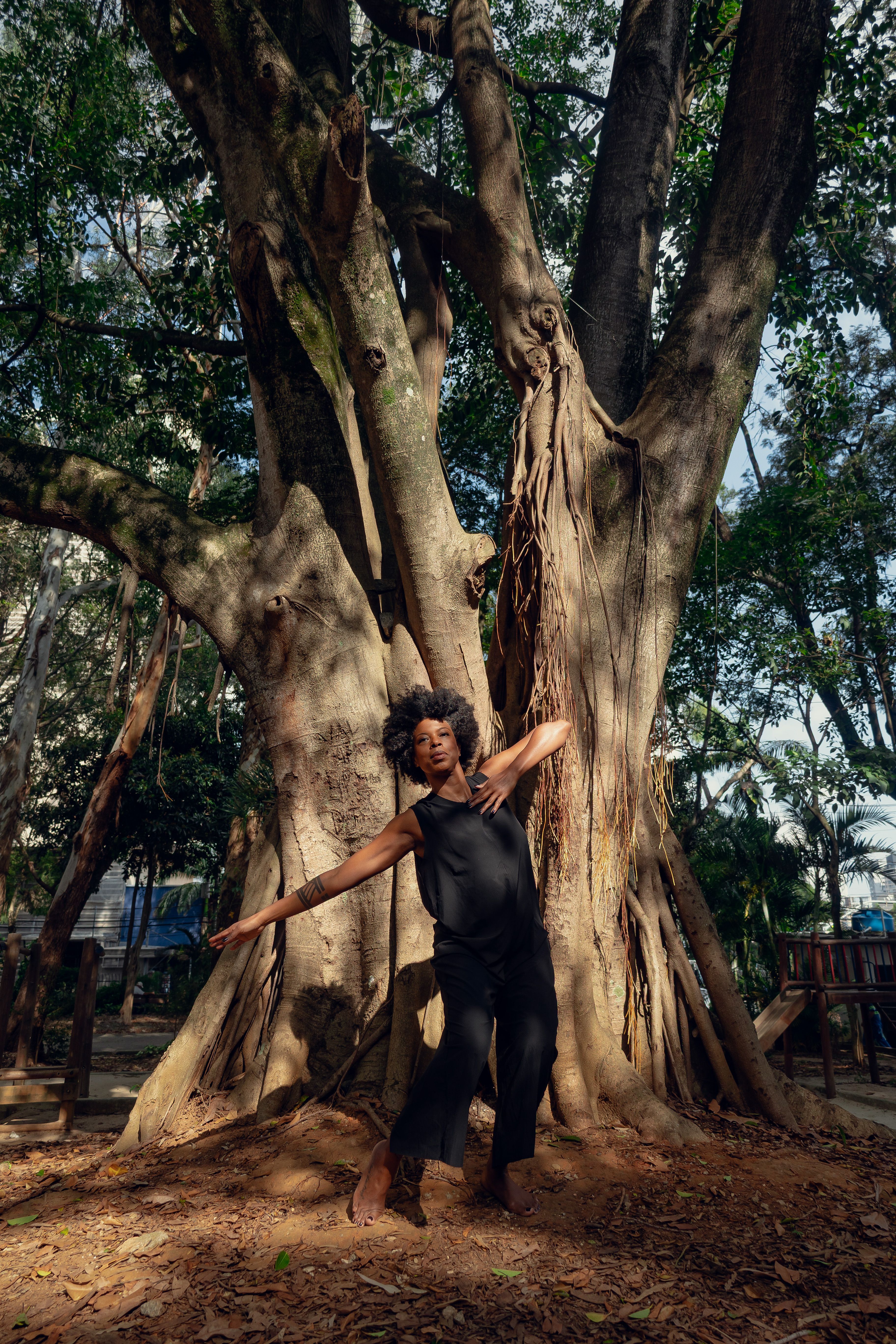 Uma mulher negra e de cabelos crespos, vestida com uma calça preta e uma regata igualmente preta, dança em frente a uma árvore de tronco largo. Ela está descalça sobre a terra e o pouco céu que se vê por trás das folhas dessa árvore é azul claro. 