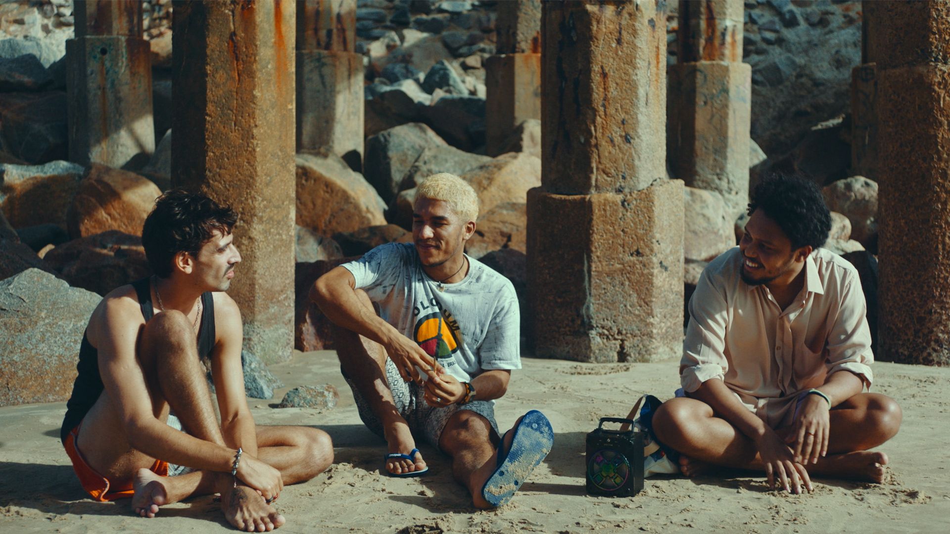 três jovens sentados na areia, conversando de forma descontraída sob uma estrutura sustentada por grossas pilastras de concreto, com grandes pedras ao fundo. O rapaz da esquerda usa regata preta e bermuda laranja; o do meio tem cabelos descoloridos e veste uma camiseta branca estampada; e o da direita, que sorri abertamente, tem cabelos crespos e veste uma camisa de botão clara. Entre eles, pousada na areia, há um rádio portátil preta, compondo uma cena casual e amigável