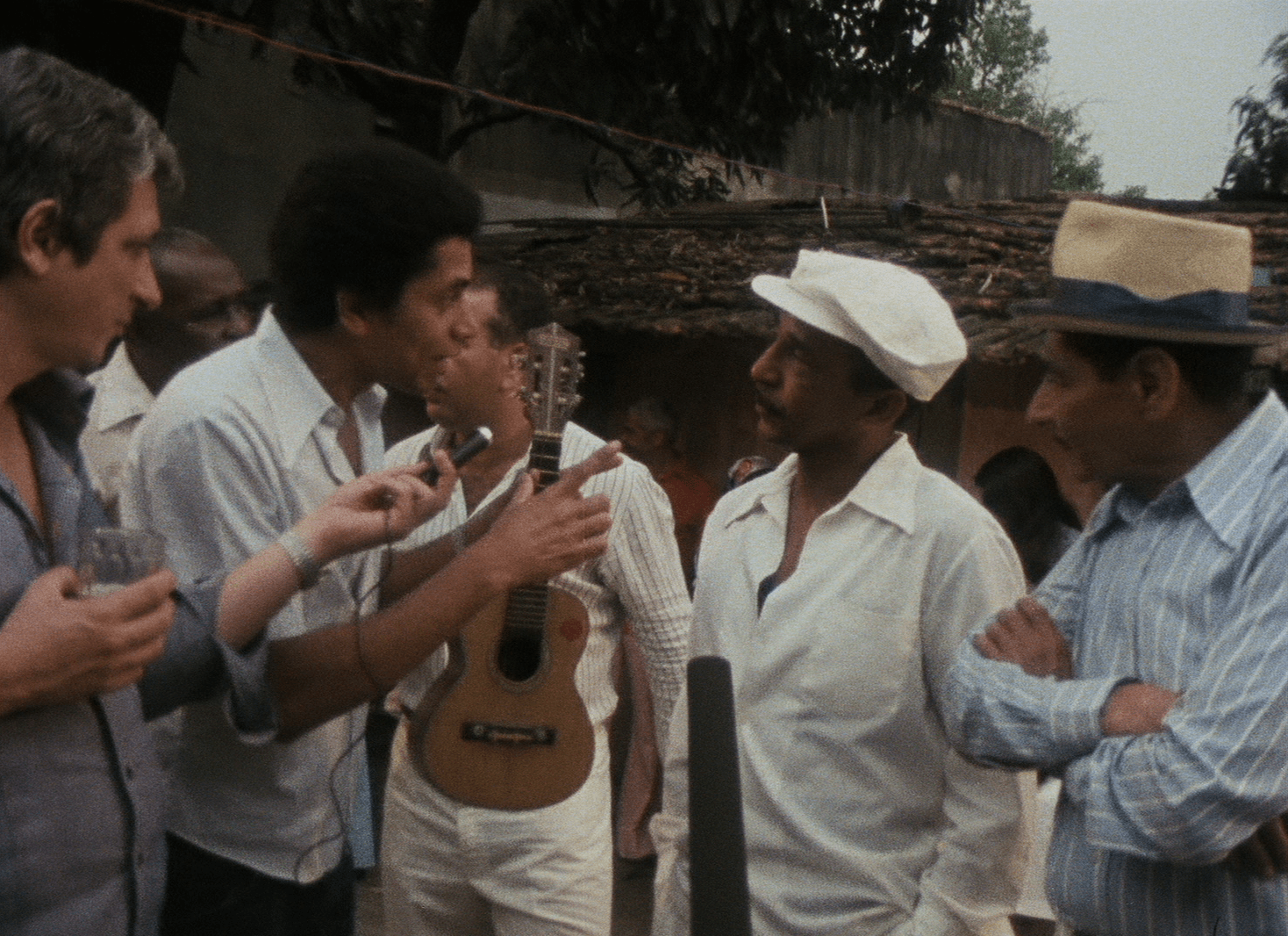 A imagem traz m&uacute;sicos conversando em uma roda, todos em p&eacute;. Muitos vestem camisas brancas ou azuis. Um deles &eacute; Paulinho da Viola.
