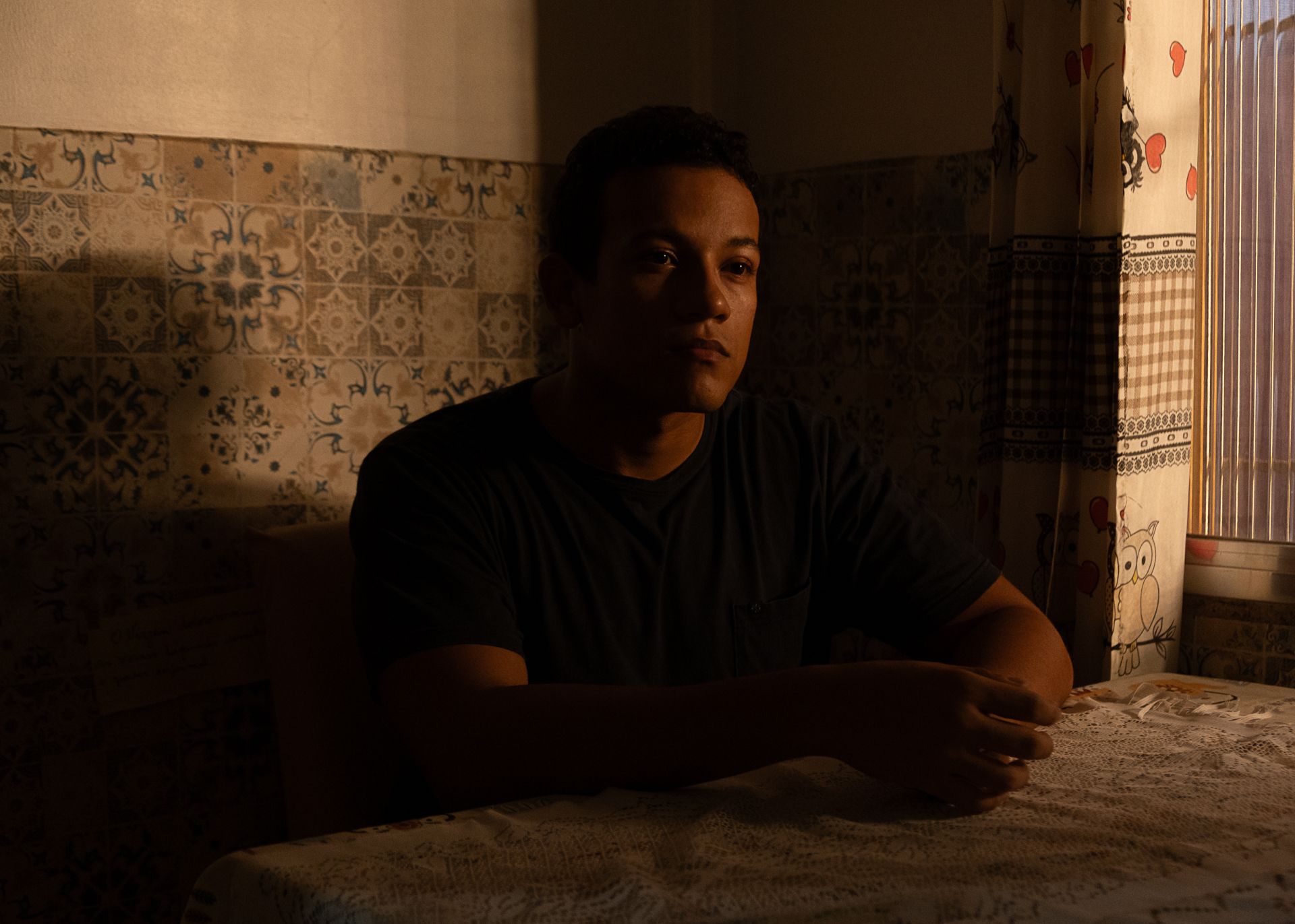 Um homem sentado &agrave; mesa de uma cozinha, iluminada pela luz que entra por uma janela &agrave; direita. O ambiente conta com azulejos decorados e cortinas estampadas, criando uma atmosfera acolhedora e dom&eacute;stica. Com express&atilde;o s&eacute;ria e olhar voltado para a janela, o sujeito transmite uma sensa&ccedil;&atilde;o de sil&ecirc;ncio, reflex&atilde;o e introspec&ccedil;&atilde;o.