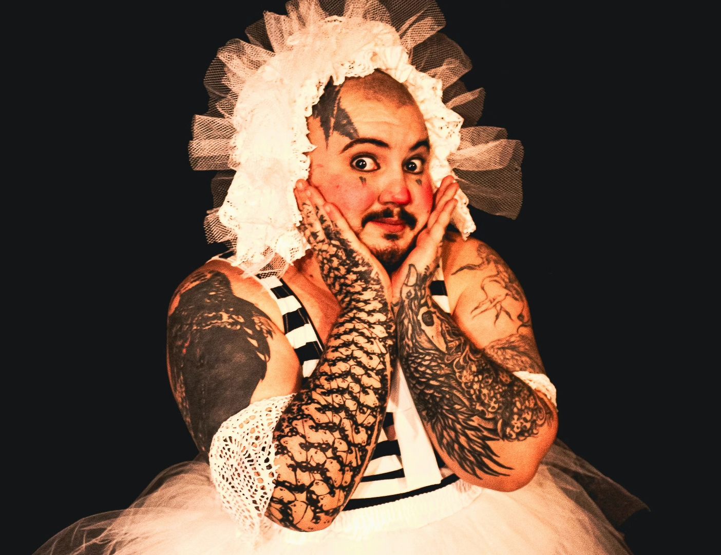 com fundo totalmente preto, retrata uma pessoa da cintura para cima em uma pose teatral e expressiva, segurando o rosto com as duas mãos e com os olhos bem arregalados. A pessoa possui cavanhaque e bigode, e usa uma maquiagem lúdica com as bochechas e a ponta do nariz avermelhadas, pequenos traços sob os olhos e uma tatuagem escura na testa. O figurino é extravagante e brinca com contrastes: na cabeça, há uma grande touca branca feita de babados de renda e tule; no tronco, veste uma peça com listras horizontais pretas e brancas acompanhada de uma saia de tule branco. Seus braços e mãos, que emolduram o rosto, são inteiramente cobertos por tatuagens escuras e detalhadas