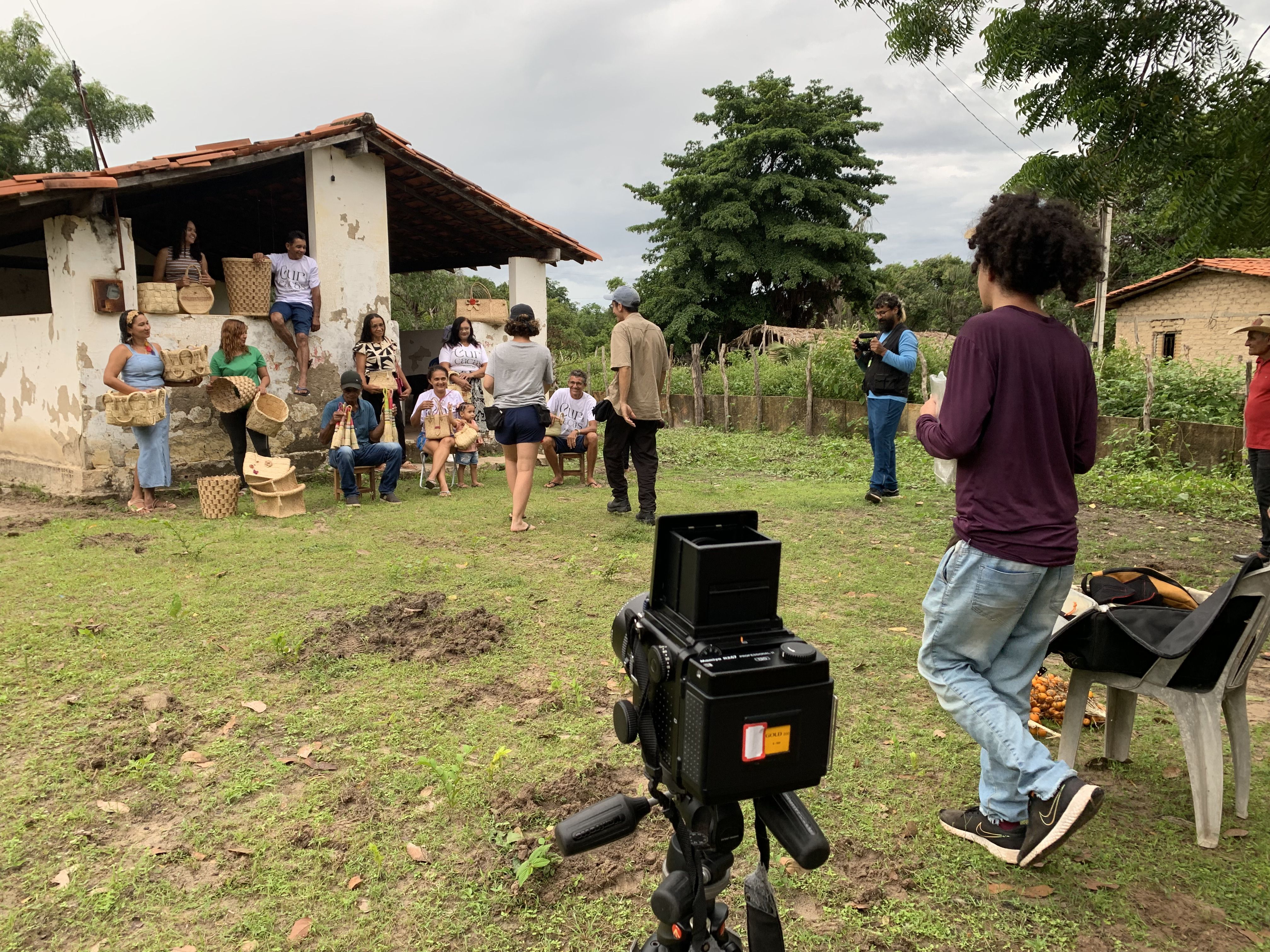 Fotografia de bastidores de uma produ&ccedil;&atilde;o audiovisual realizada em &aacute;rea rural. Em frente a uma casa simples, um grupo de homens, mulheres e crian&ccedil;as posa com pe&ccedil;as de artesanato em fibra vegetal, enquanto uma c&acirc;mera fotogr&aacute;fica e integrantes da equipe aparecem em primeiro plano. Ao fundo, &aacute;rvores, cercas e um c&eacute;u nublado comp&otilde;em a paisagem do campo.