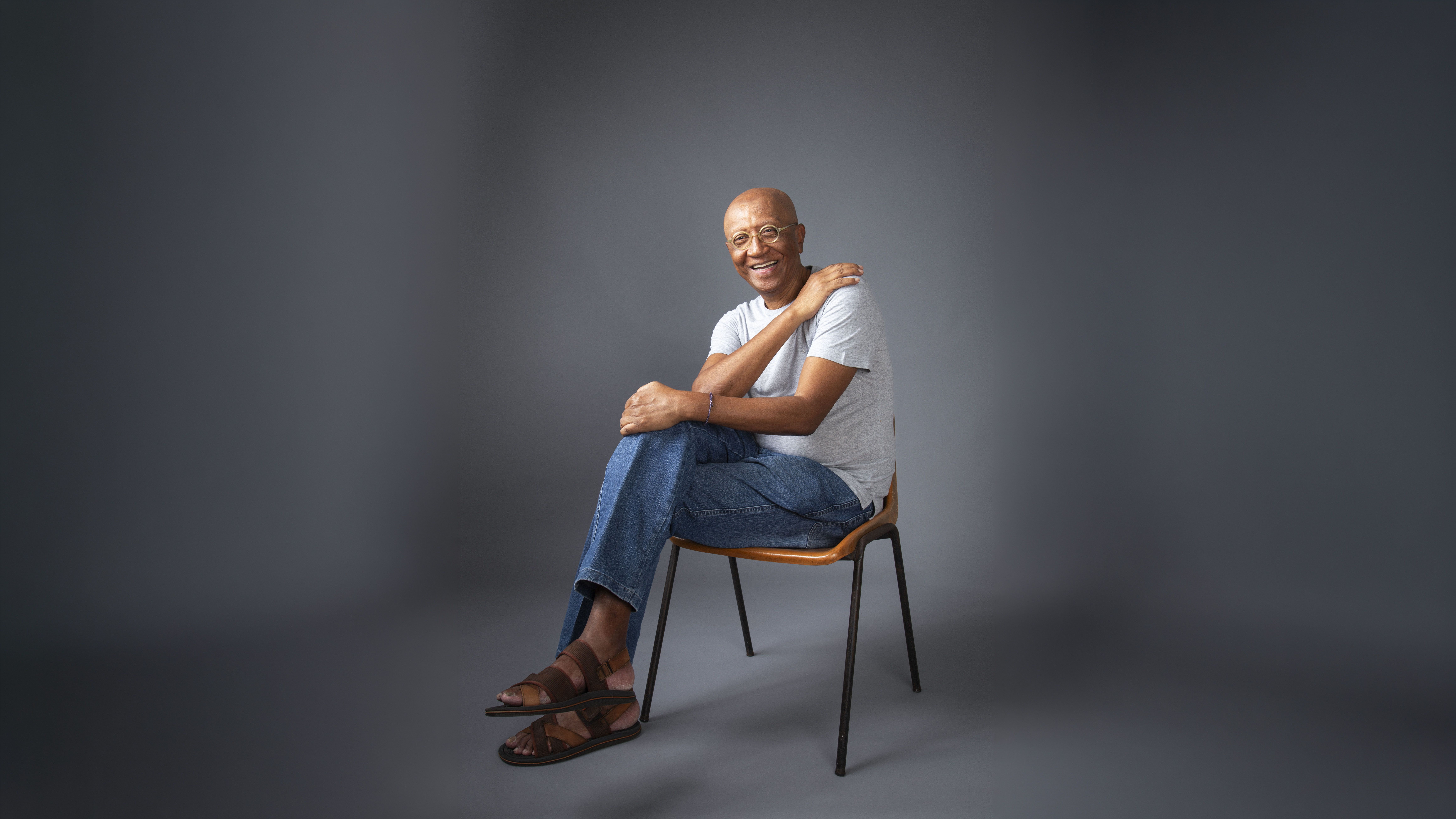 Retrato horizontal de Helio Santos, um homem negro de pele clara e careca, sentado e sorrindo. Ele usa óculos redondos, camiseta cinza, calça jeans azul e sandálias de couro marrom. Suas pernas estão cruzadas e a mão esquerda pousa sobre o ombro direito. Ele está sentado em uma cadeira simples com assento de madeira e pernas de metal. O fundo é cinza texturizado e a iluminação suave e quente vem da esquerda. Retrato horizontal de Helio Santos, um homem negro de pele clara e careca, sentado e sorrindo. Ele usa óculos redondos, camiseta cinza, calça jeans azul e sandálias de couro marrom. Suas pernas estão cruzadas e a mão esquerda pousa sobre o ombro direito. Ele está sentado em uma cadeira simples com assento de madeira e pernas de metal. O fundo é cinza texturizado e a iluminação suave e quente vem da esquerda.