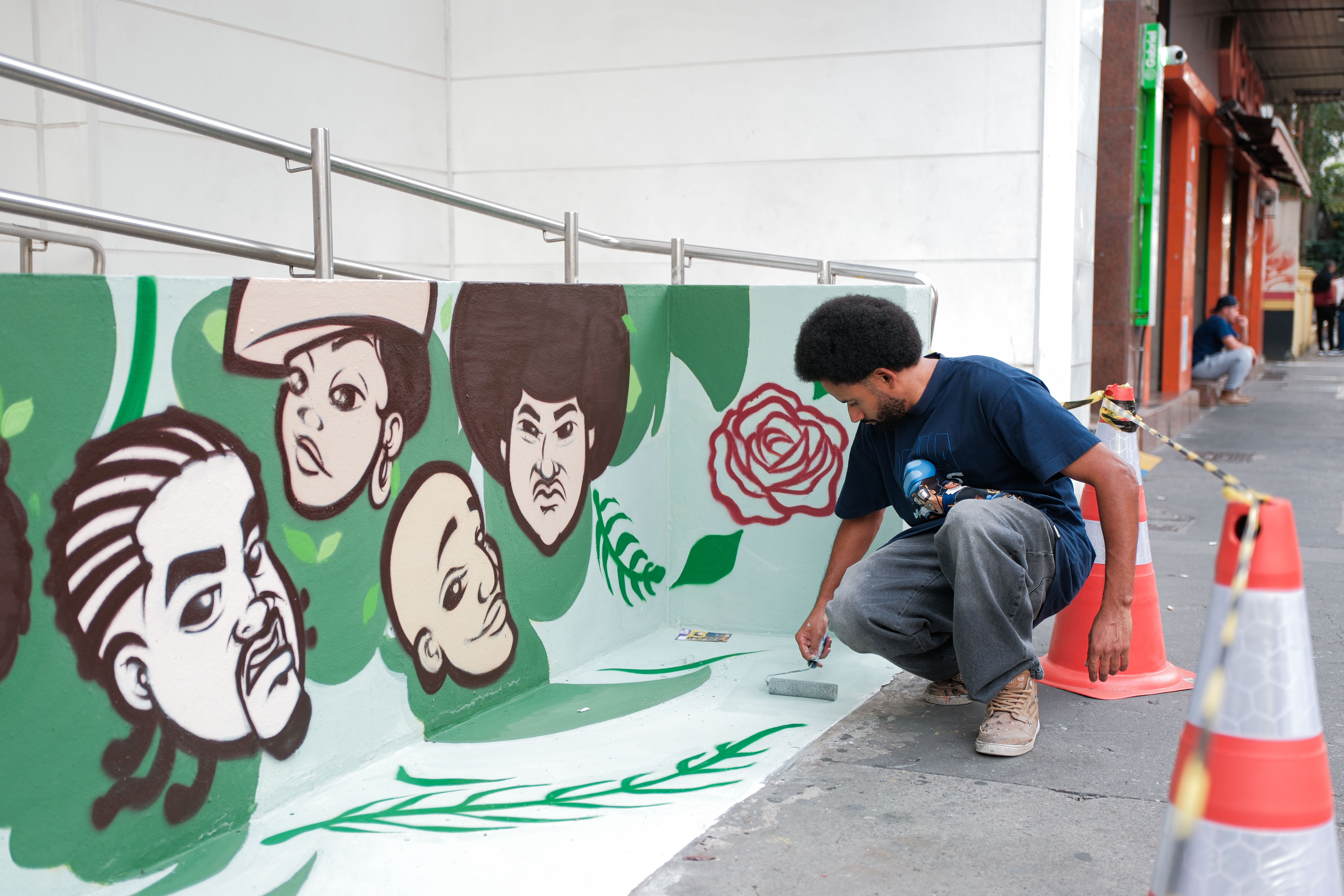 Ricardo Sotaq, de cabelos crespos e barba, agachado na calçada enquanto trabalha na pintura do banco de concreto. Ele utiliza um pequeno rolo de espuma para pintar o assento. O encosto do banco já exibe grande parte do mural: uma composição de rostos estilizados em tons de marrom escuro e branco, com variados traços e tipos de cabelo, flutuando sobre um fundo verde decorado com formas de folhas e o contorno vermelho de uma rosa. O espaço de trabalho do artista está protegido por cones de trânsito laranjas com fita de isolamento preta e amarela, separando-o do movimento de pedestres na rua ao fundo.