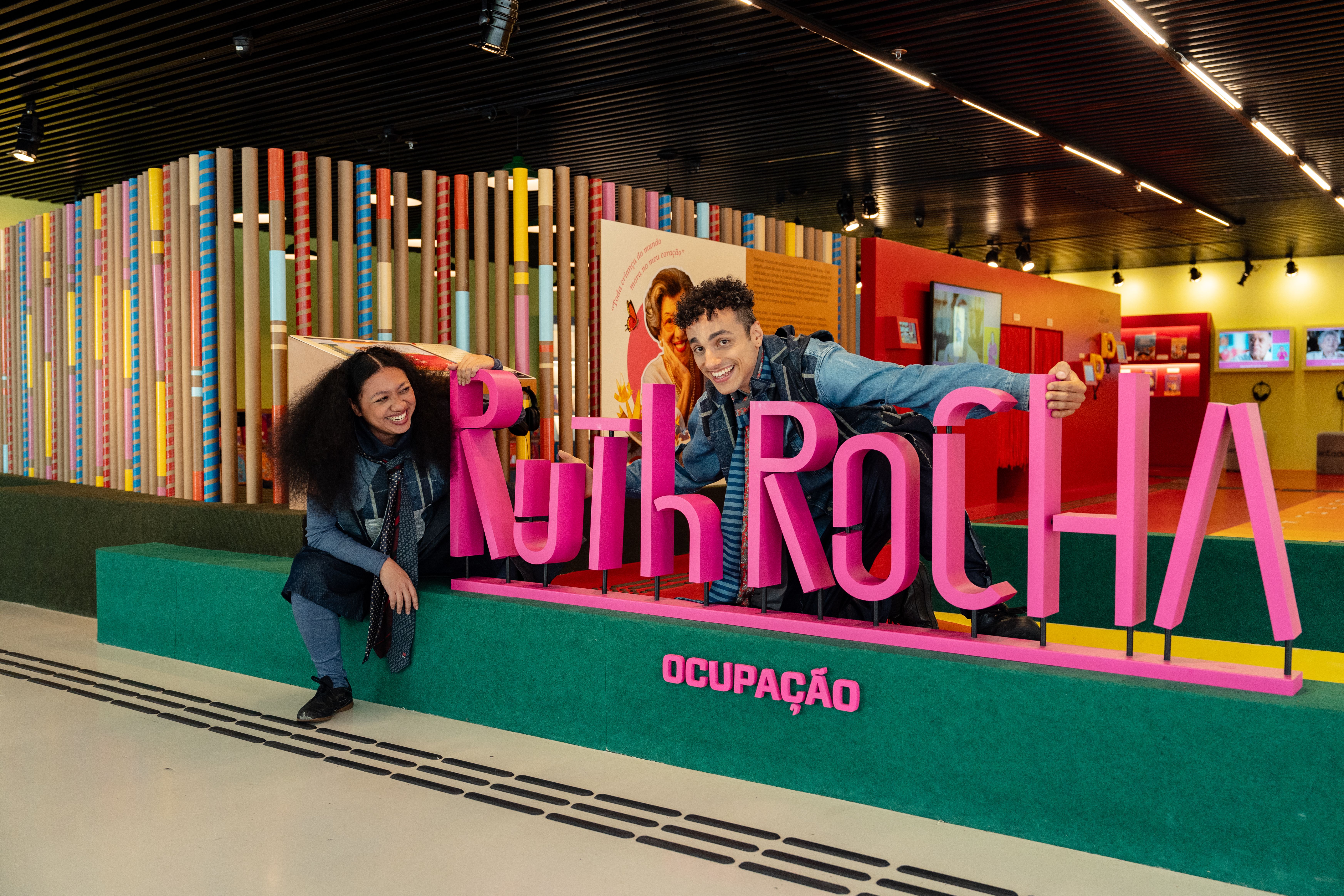 A fotografia mostra duas pessoas interagindo com um grande letreiro rosa tridimensional escrito "Ruth RocHA", apoiado sobre uma base verde retangular que traz a palavra "OCUPAÇÃO" também em rosa. O cenário é o interior de uma exposição iluminada e colorida. Ao fundo, há uma divisória feita com longos tubos de papelão coloridos e uma parede vermelha com telas exibindo vídeos. O chão claro possui linhas de piso tátil para acessibilidade. À esquerda, uma mulher de cabelos escuros e volumosos, vestindo uma sobreposição de roupas em tons de azul e cinza com um lenço no pescoço, está sentada na base verde olhando para o lado com uma expressão feliz. À direita, um jovem sorridente de jaqueta jeans debruça-se entre as palavras do letreiro, apoiando o rosto na mão enquanto olha para a câmera.