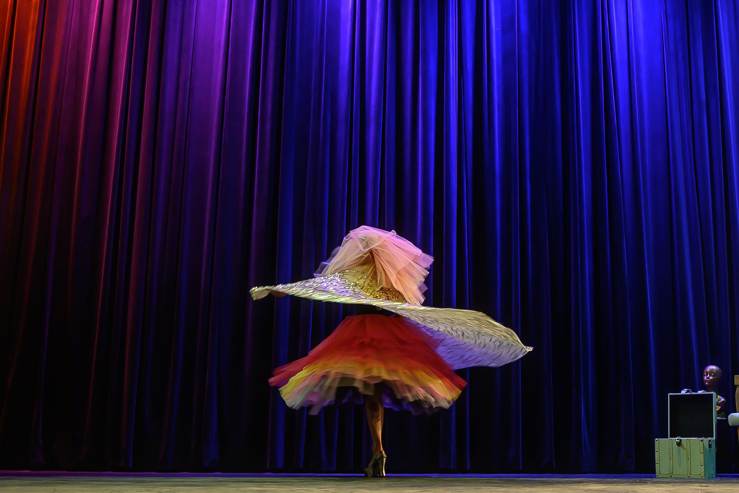 Uma dançarina de costas é fotografada em um giro veloz no palco. Ela veste um tutu de tule em degradê colorido (vermelho, amarelo e rosa), salto plataforma dourado e segura grandes asas laminadas brilhantes. O palco preto tem como fundo cortinas de veludo em um gradiente que vai do laranja ao azul-real. No canto inferior direito, avistam-se uma maleta verde-água e um busto manequim metálico sob iluminação cênica.