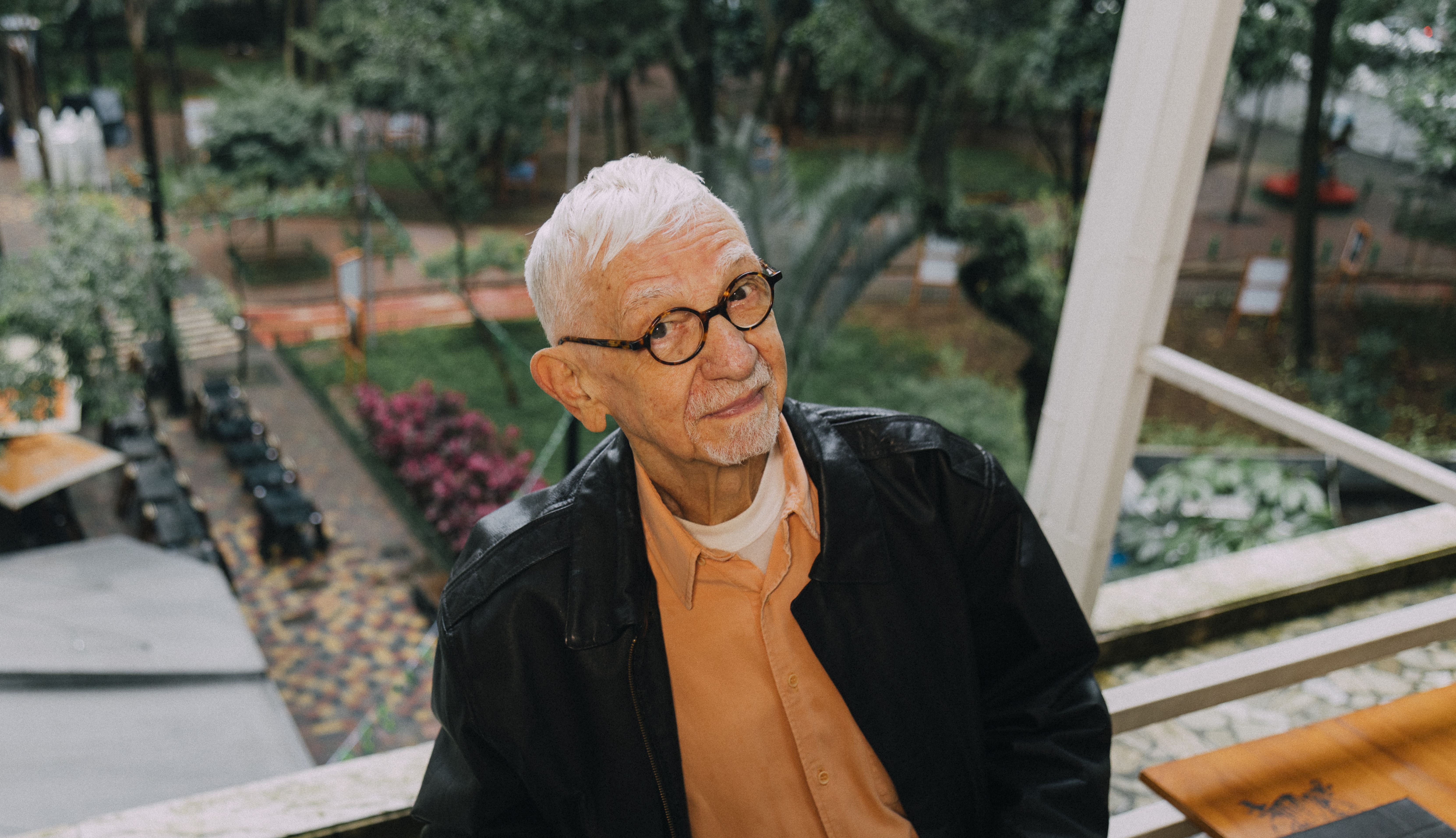 João Silvério Trevisan é um senhor branco, com cabelos brancos e óculos de grau. Ele veste uma blusa laranja e uma jaqueta preta. Ao lado e ao fundo dele, há mesas e cadeiras de madeira.