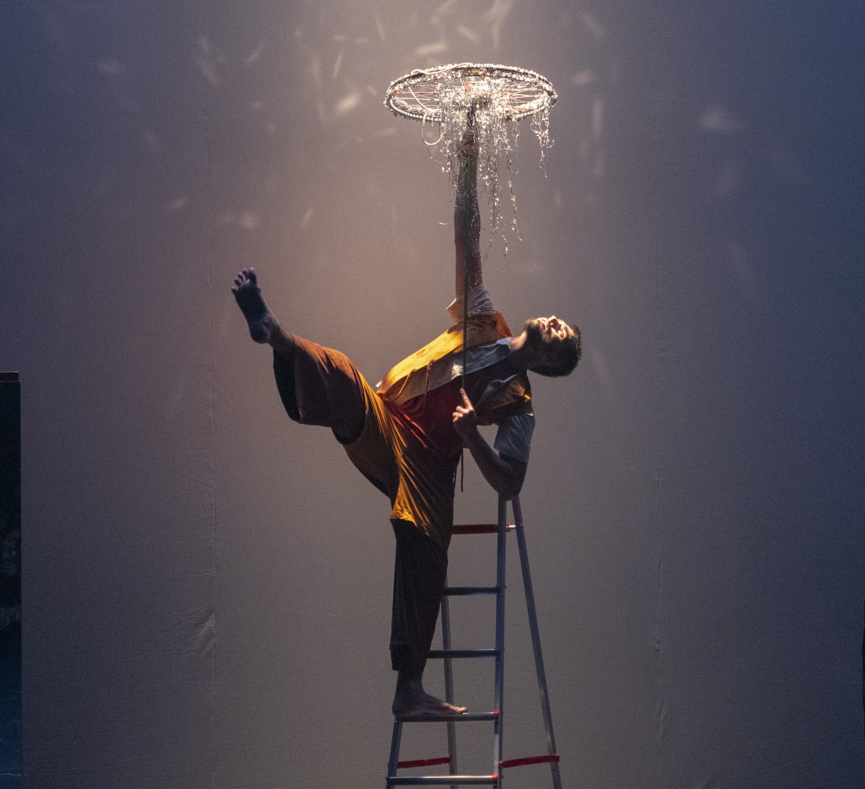 oto colorida, em plano inteiro e enquadramento centralizado, capturando uma performance art&iacute;stica em um palco. Um homem barbudo est&aacute; equilibrado no topo de uma escada de alum&iacute;nio aberta. Ele veste cal&ccedil;a larga marrom e uma blusa em tons de amarelo e laranja com mangas brancas. O artista realiza uma pose acrob&aacute;tica: seu p&eacute; esquerdo apoia-se em um dos degraus superiores, enquanto sua perna direita est&aacute; esticada para o alto e para o lado esquerdo, com o p&eacute; descal&ccedil;o apontado para cima. Com o bra&ccedil;o esquerdo estendido verticalmente, ele equilibra uma estrutura circular prateada, semelhante a uma roda de bicicleta decorada, da qual caem fios brilhantes e franjas prateadas como se fosse uma lumin&aacute;ria ou guarda-chuva de fitas. A ilumina&ccedil;&atilde;o &eacute; c&ecirc;nica e dram&aacute;tica, com um feixe de luz focado de cima para baixo diretamente sobre o objeto prateado, criando reflexos pontuais e texturas luminosas contra o fundo cinza-azulado e enevoado do palco.