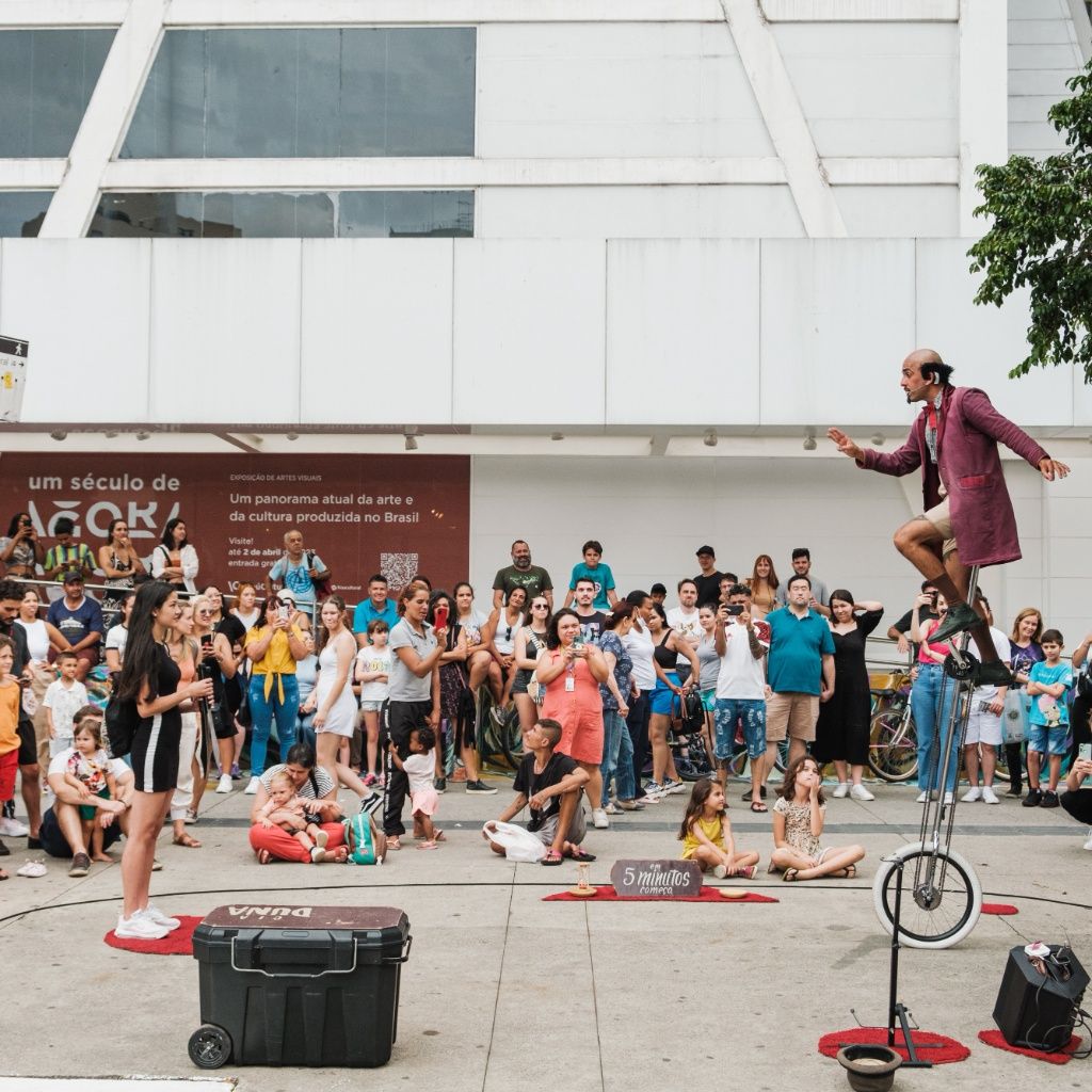Foto colorida, em plano aberto e enquadramento centralizado, capturando uma apresenta&ccedil;&atilde;o art&iacute;stica de rua em uma pra&ccedil;a p&uacute;blica. Do lado direito, um homem calvo com roupas perform&aacute;ticas &mdash; casaco longo de tom magenta ou vinho, bermuda clara e meias altas &mdash; equilibra-se no topo de um monociclo alto, mantendo os bra&ccedil;os abertos para os lados. Ele olha em dire&ccedil;&atilde;o ao lado esquerdo da imagem, onde uma jovem de pele clara, cabelos longos e escuros e vestido preto curto o observa de p&eacute; com um microfone na m&atilde;o.

Ao redor deles, dezenas de espectadores de idades variadas formam uma grande plateia semicircular. Algumas pessoas est&atilde;o sentadas no ch&atilde;o, incluindo crian&ccedil;as e uma m&atilde;e com um beb&ecirc;, enquanto a maioria permanece de p&eacute; registrando a cena com celulares. No ch&atilde;o da pra&ccedil;a, h&aacute; equipamentos de apoio do artista: uma grande caixa preta com rodinhas no canto inferior esquerdo, uma pequena placa de madeira onde se l&ecirc; escrito &agrave; m&atilde;o 