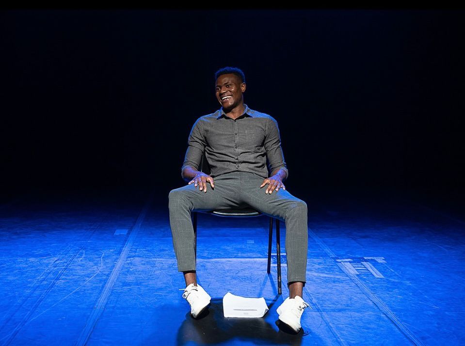 Manuel Kafina est&aacute; sorrindo sentado em uma cadeira sobre um palco azulado. Ele &eacute; um homem negro, veste cal&ccedil;a e camisa social cinzas combinadas com t&ecirc;nis brancos. H&aacute; um roteiro ca&iacute;do no ch&atilde;o entre seus p&eacute;s.