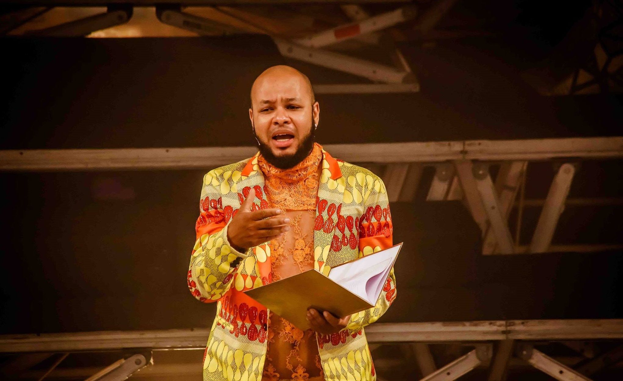 Nando Z&acirc;mbia apresenta-se, de forma expressiva, em um palco. Ele &eacute; um homem negro e calvo, veste um blazer com estampas vibrantes em amarelo e vermelho sobre uma blusa de renda. O autor segura uma pasta aberta.