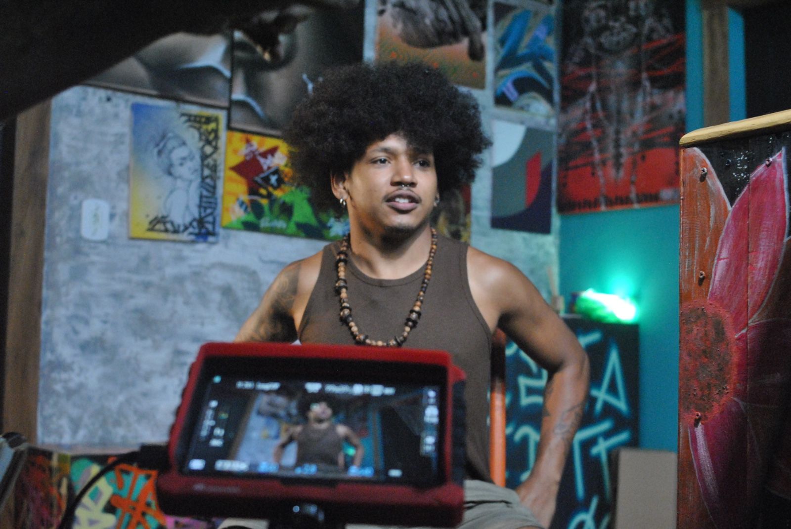 Um homem negro, com colar de contas, posa ao fundo com as m&atilde;os na cintura. Em primeiro plano, um monitor de grava&ccedil;&atilde;o com capa vermelha exibe e enquadra a imagem do homem. O cen&aacute;rio &eacute; decorado com diversas artes urbanas, pinturas coloridas e uma parede texturizada de cimento.