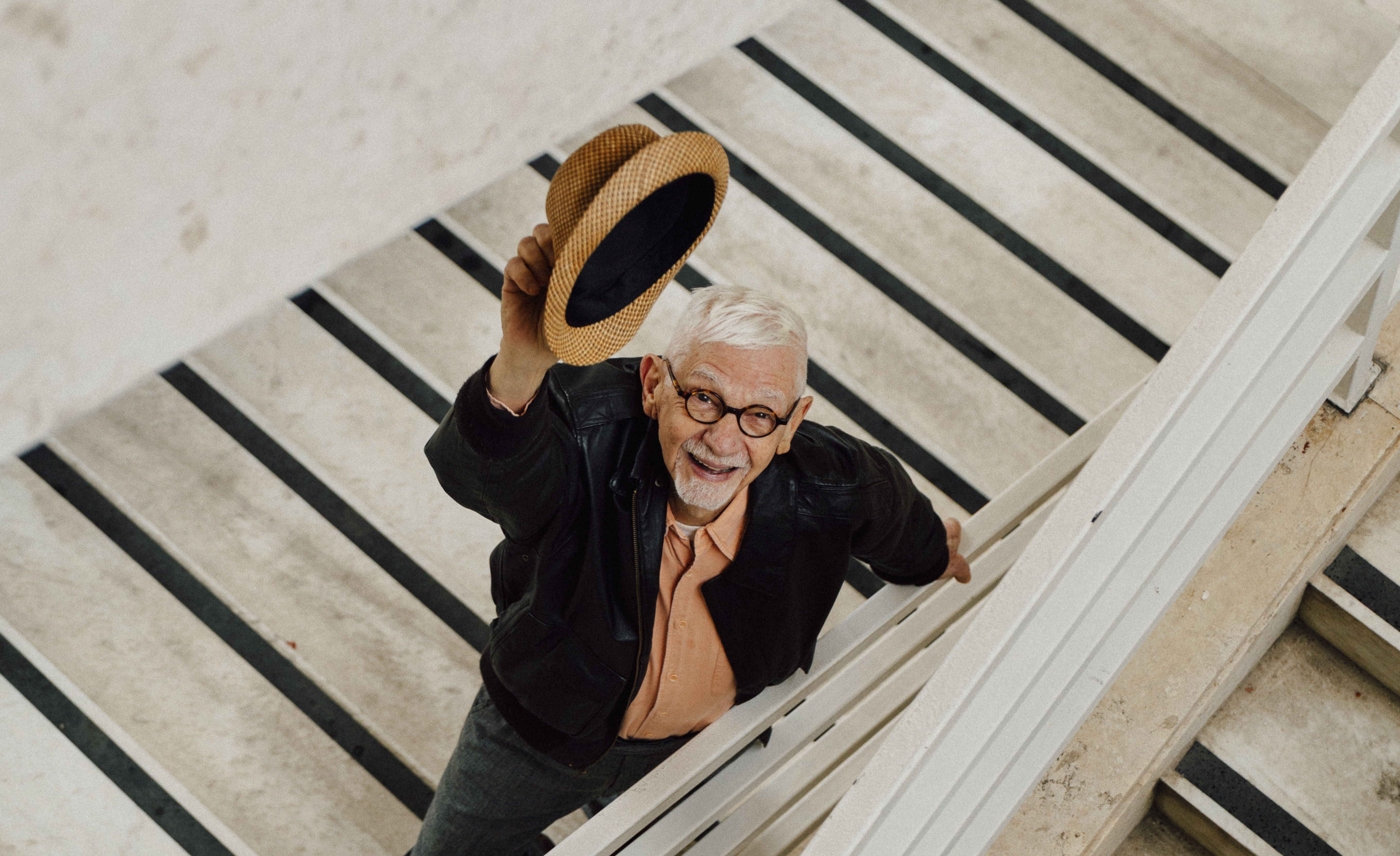 João Silvério Trevisan, um homem branco e com cabelos brancos, sorri. Ele usa óculos de grau e olha para cima em uma escadaria. Ele veste uma jaqueta preta sobre uma camisa salmão e ergue um chapéu de palha com a mão direita. A foto foi tirada de um ângulo superior, destacando as linhas diagonais dos degraus e do corrimão. João Silvério Trevisan, um homem branco e com cabelos brancos, sorri. Ele usa óculos de grau e olha para cima em uma escadaria. Ele veste uma jaqueta preta sobre uma camisa salmão e ergue um chapéu de palha com a mão direita. A foto foi tirada de um ângulo superior, destacando as linhas diagonais dos degraus e do corrimão.