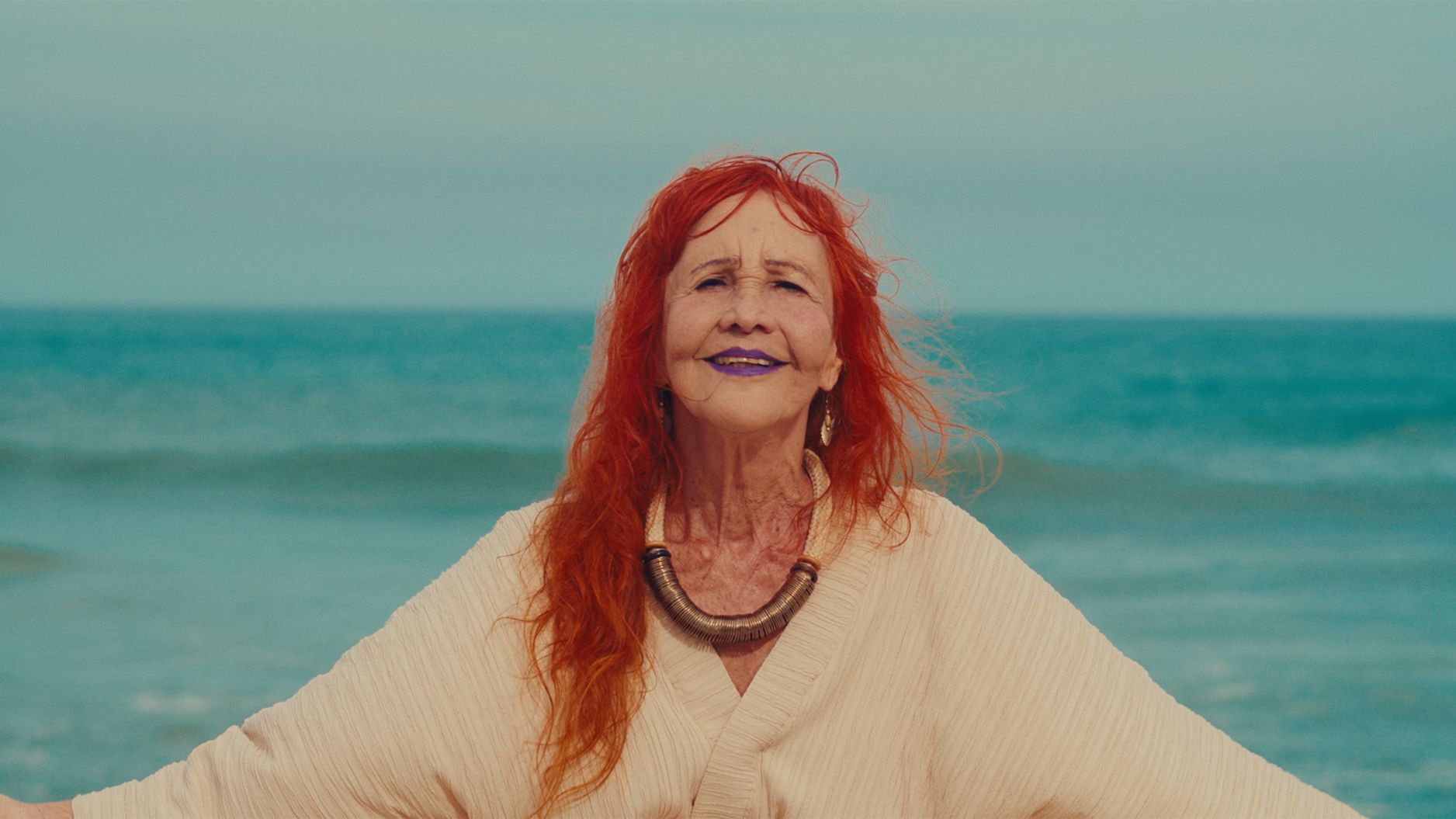 A imagem mostra uma mulher sorridente, com cabelos longos e ruivos e batom roxo. Ela está posicionada no centro, de braços abertos, vestindo uma roupa clara e um colar rústico volumoso. O fundo é composto por uma praia, destacando o azul do mar e um céu em tons semelhantes. Ela tem uma expressão alegre. A imagem mostra uma mulher sorridente, com cabelos longos e ruivos e batom roxo. Ela está posicionada no centro, de braços abertos, vestindo uma roupa clara e um colar rústico volumoso. O fundo é composto por uma praia, destacando o azul do mar e um céu em tons semelhantes. Ela tem uma expressão alegre.