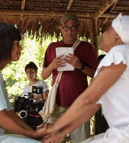 A imagem mostra duas mulheres de destaque vestindo roupa branca. A da esquerda tem apar&ecirc;ncia mais jovem e est&aacute; segurando a m&atilde;o da mulher da direita, que aparenta ser um mais velha e uma lideran&ccedil;a espiritual. Ao fundo, acompanhando a cena, est&aacute; o diretor e produtor Silvio Margarido e, atr&aacute;s dele, uma cinegrafista que registra a cena