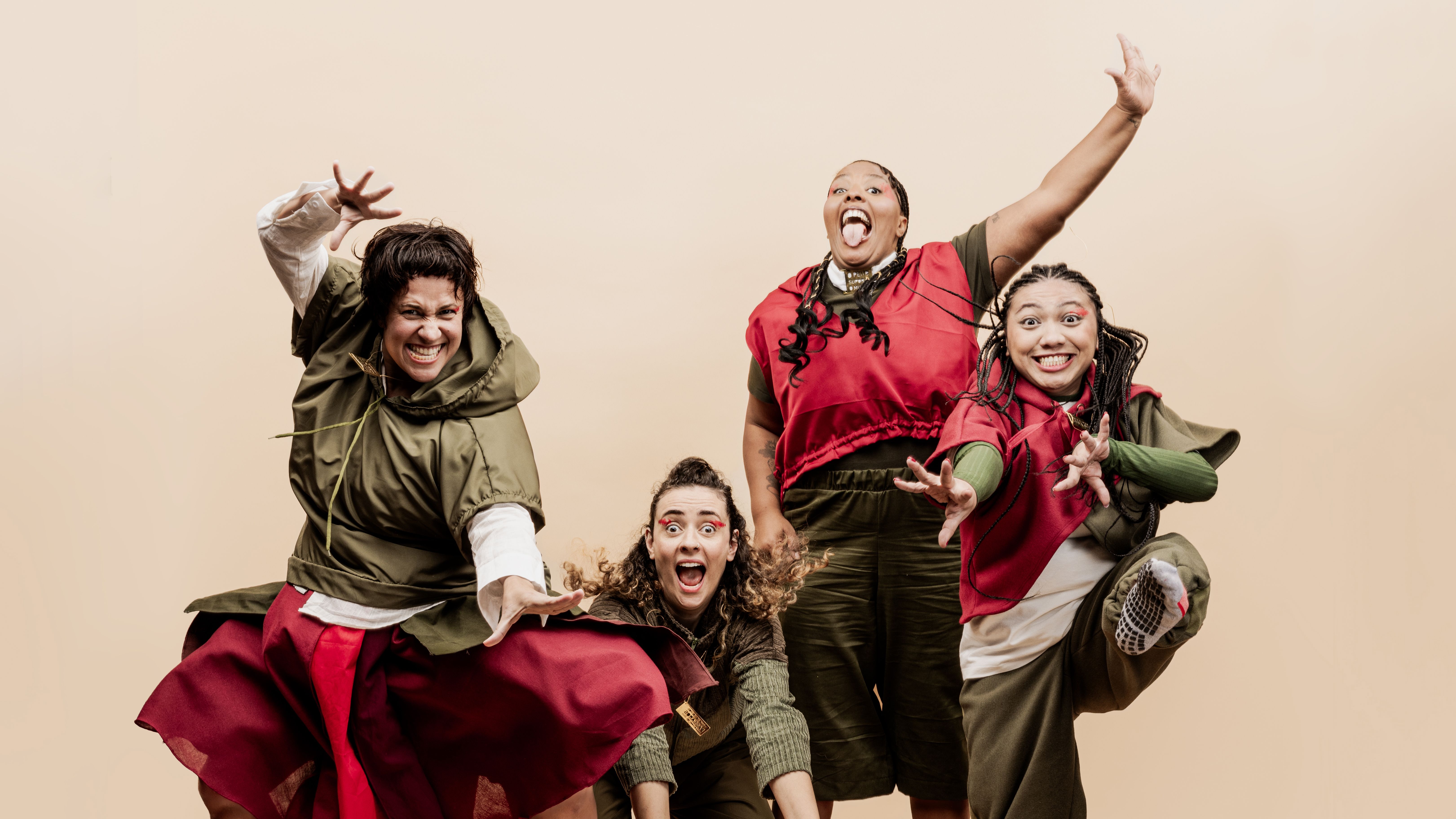 Fotografia colorida com fundo bege plano. Quatro pessoas fazem poses expressivas e divertidas. Todas usam maquiagem artística vermelha nos olhos e figurinos em tons de verde-militar e vermelho.

À esquerda, uma mulher de cabelo curto e capuz verde faz uma pose forte de salto, estendendo as mãos com os dedos curvos como garras. Ao centro, uma mulher de cabelos cacheados sorri de boca aberta, abaixada. Atrás dela, uma mulher negra com tranças comemora alegremente com a língua para fora e um braço erguido. À direita, uma mulher com tranças finas equilibra-se em uma perna só, sorrindo e projetando as mãos à frente.