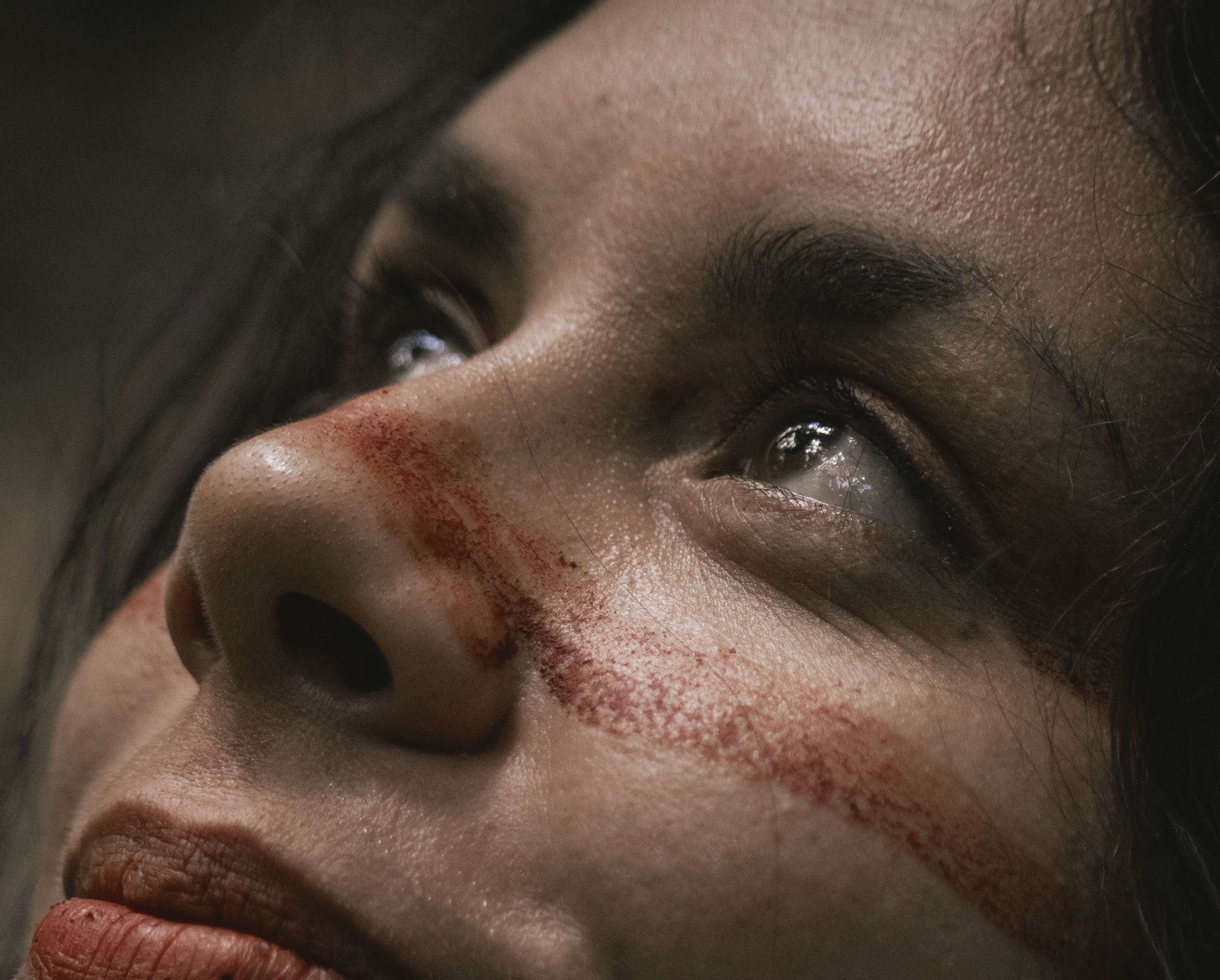A fotografia captura o rosto de uma mulher indígena com expressão serena e um leve sorriso, olhando para o alto com os olhos brilhantes refletindo a luz. Sua pele apresenta textura natural, com poros e pequenas pintas visíveis, e está adornada por pinturas faciais tradicionais na cor vermelho-terra: um traço horizontal espesso que atravessa as bochechas e a ponte do nariz, além de marcações verticais que descem pelo queixo. Com os cabelos escuros caindo levemente pelas laterais, a imagem transmite uma sensação de ancestralidade, força e contemplação.