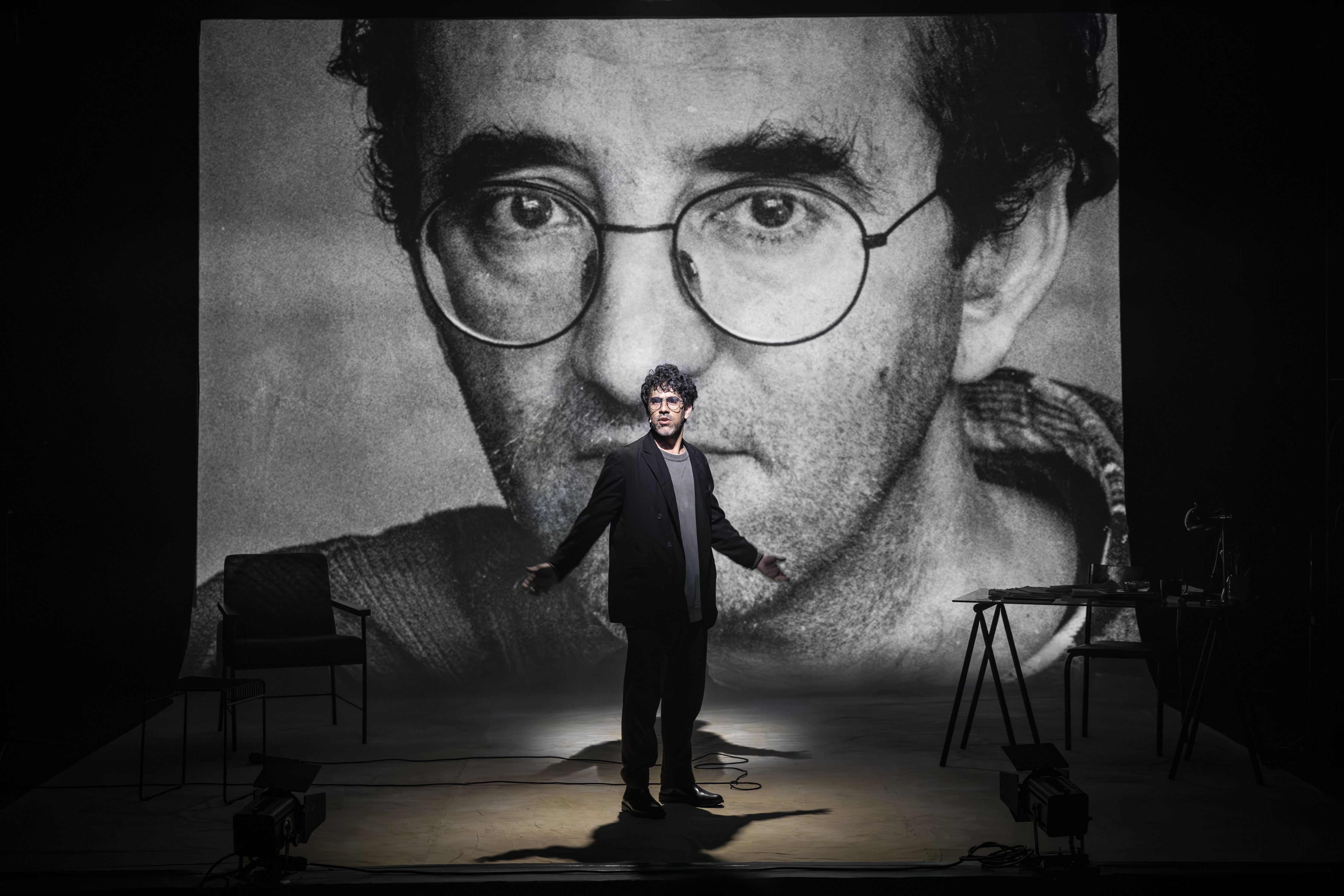Fotografia na horizontal de um palco teatral iluminado no centro. Em primeiro plano, um homem de cabelos cacheados e óculos de grau veste blazer preto sobre camiseta cinza e calça escura. Ele está de pé, com os braços abertos e as palmas das mãos voltadas para cima. Ao fundo, uma grande projeção em preto e branco exibe o rosto do escritor Roberto Bolaño em close-up, olhando fixamente para a frente com óculos redondos. O cenário ao redor inclui uma cadeira à esquerda e uma mesa com cadeira à direita, sob fundo escuro, destacando a figura central sob um foco de luz no chão. Fotografia na horizontal de um palco teatral iluminado no centro. Em primeiro plano, um homem de cabelos cacheados e óculos de grau veste blazer preto sobre camiseta cinza e calça escura. Ele está de pé, com os braços abertos e as palmas das mãos voltadas para cima. Ao fundo, uma grande projeção em preto e branco exibe o rosto do escritor Roberto Bolaño em close-up, olhando fixamente para a frente com óculos redondos. O cenário ao redor inclui uma cadeira à esquerda e uma mesa com cadeira à direita, sob fundo escuro, destacando a figura central sob um foco de luz no chão.