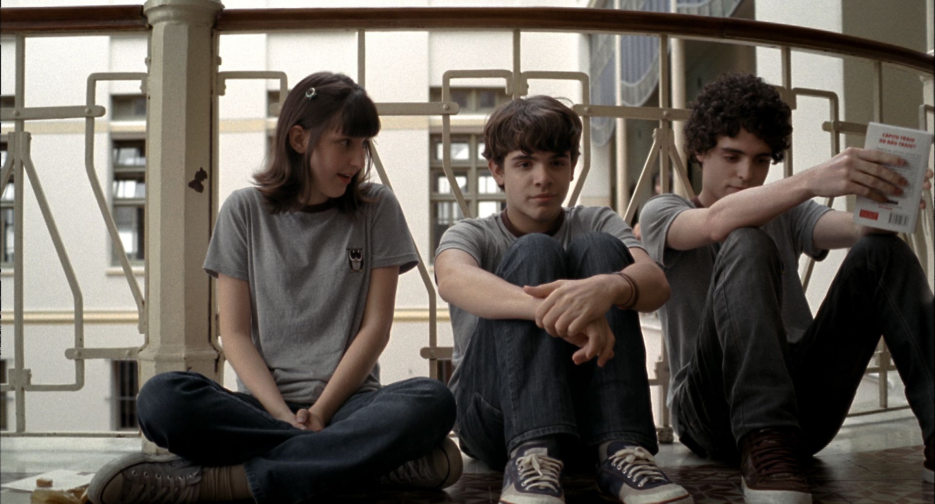 Três adolescentes brancos sentados no chão de uma varanda com grades geométricas. A menina e os meninos usam camisetas cinzas e calças jeans, conversando à luz do dia. Três adolescentes brancos sentados no chão de uma varanda com grades geométricas. A menina e os meninos usam camisetas cinzas e calças jeans, conversando à luz do dia.