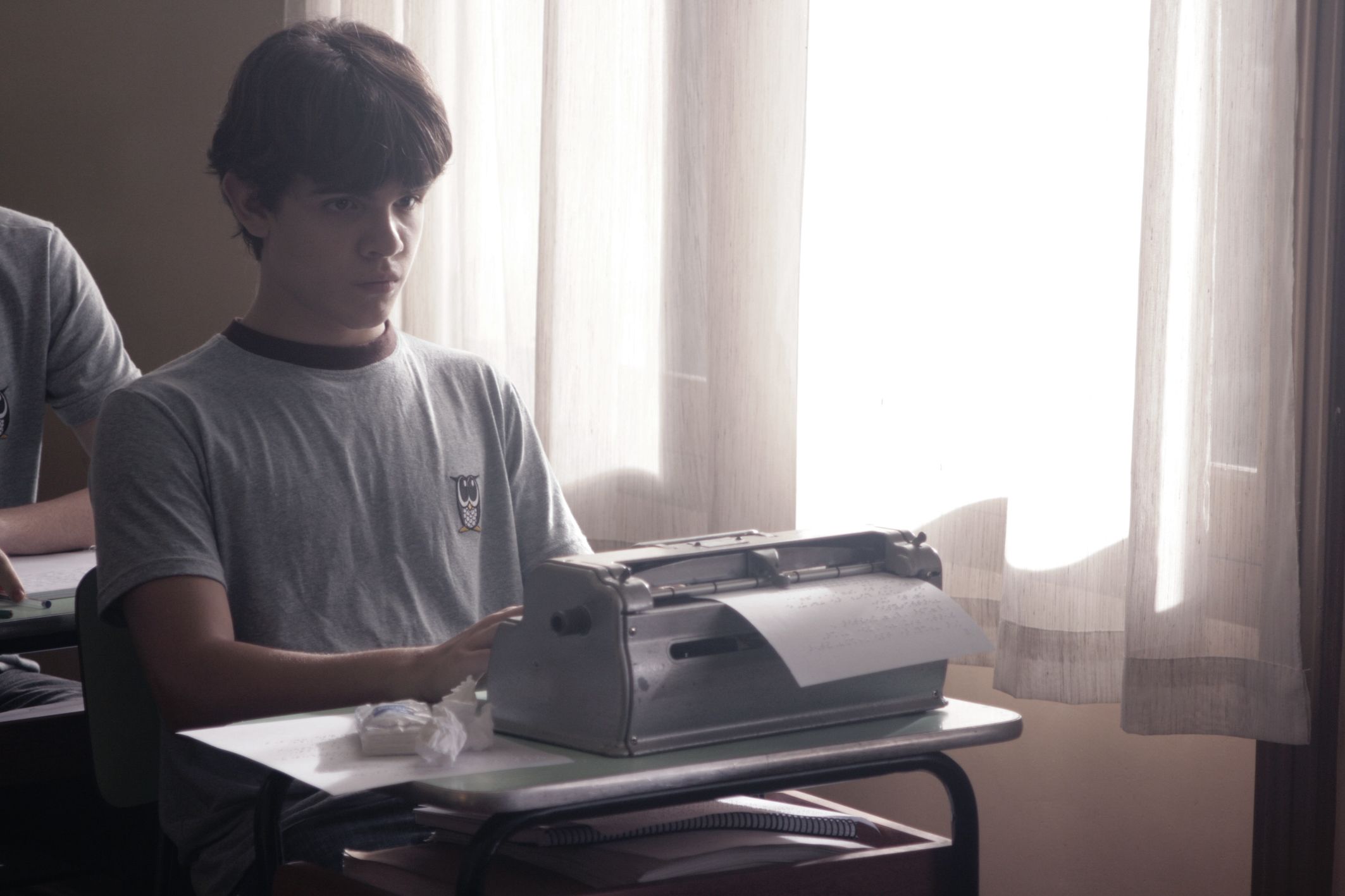 Fotografia colorida de menino branco de cabelos castanhos, sentado diante de uma m&aacute;quina de escrever cinza sobre mesa. Ele usa camiseta cinza. Ao fundo, janela com cortina branca.