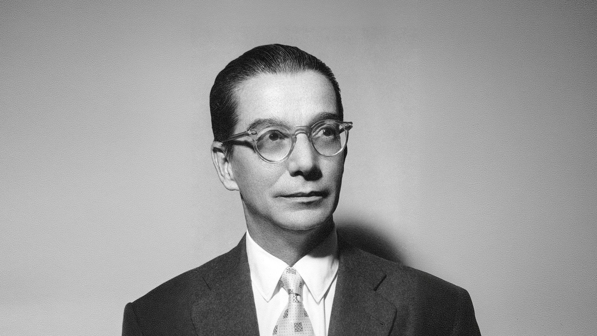 Fotografia em preto e branco de Anísio, um homem de meia-idade, em plano de busto. Ele veste terno escuro, camisa branca e gravata estampada. Usa óculos de armação transparente e tem o cabelo escuro, curto e bem penteado para trás. Seu rosto está virado para a direita, olhando para fora da cena com expressão séria. O fundo é uma parede cinza texturizada e uniforme.
