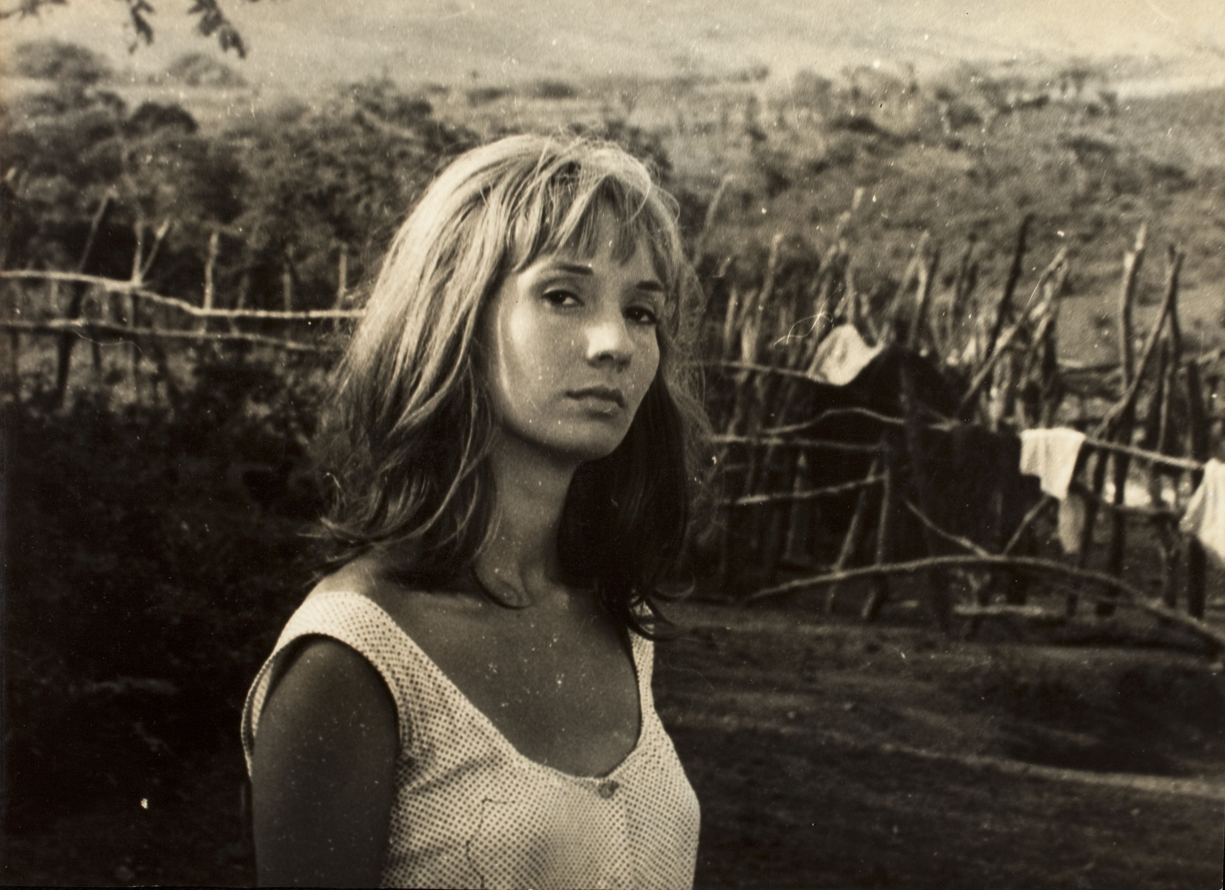 Helena Ignez aparece em primeiro plano, com expressão séria e contemplativa. A fotografia em preto e branco mostra a atriz em um ambiente rural, onde há uma cerca e vegetação. Ao fundo, roupas penduradas e a paisagem campestre reforçam a atmosfera simples e cotidiana da cena. Helena Ignez aparece em primeiro plano, com expressão séria e contemplativa. A fotografia em preto e branco mostra a atriz em um ambiente rural, onde há uma cerca e vegetação. Ao fundo, roupas penduradas e a paisagem campestre reforçam a atmosfera simples e cotidiana da cena.
