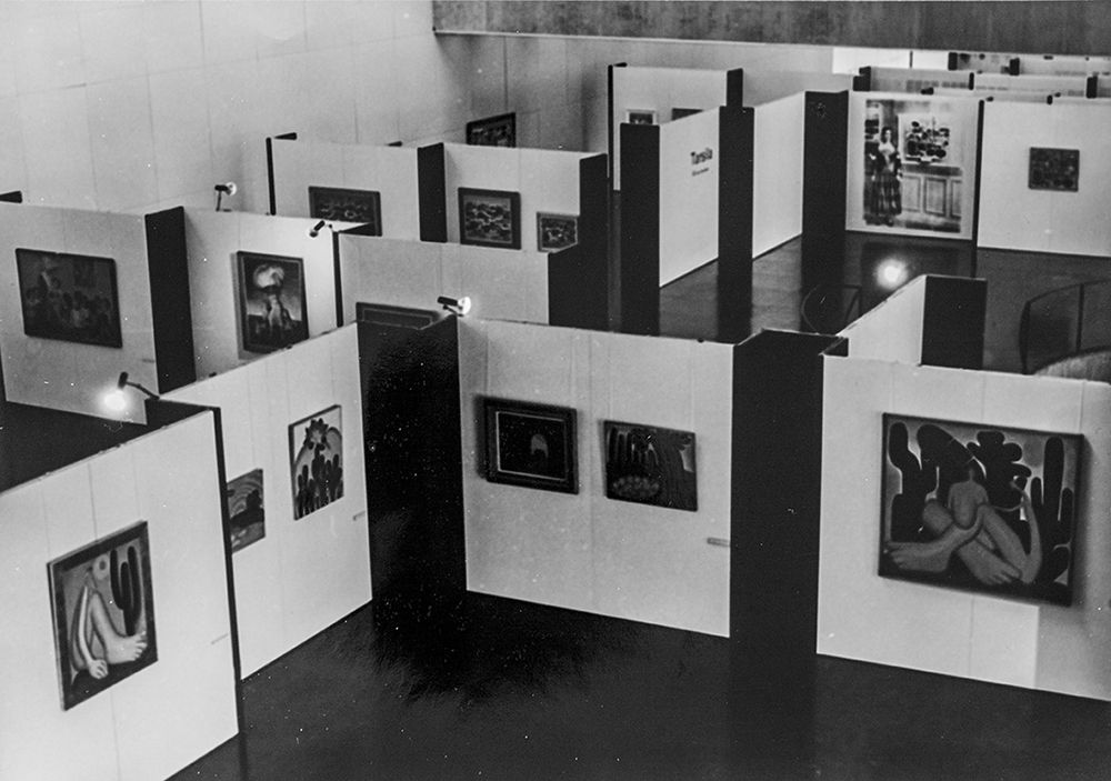Fotografia em preto e branco com vista a&eacute;rea de uma exposi&ccedil;&atilde;o de arte. No centro, diversos pain&eacute;is expostos formam divis&oacute;rias e corredores geom&eacute;tricos sobre um piso escuro. Presas a esses pain&eacute;is claros, h&aacute; v&aacute;rias quadros e fotografias emoldurados, distribu&iacute;dos ao longo do espa&ccedil;o.

Na parte inferior direita, destaca-se o famoso quadro 