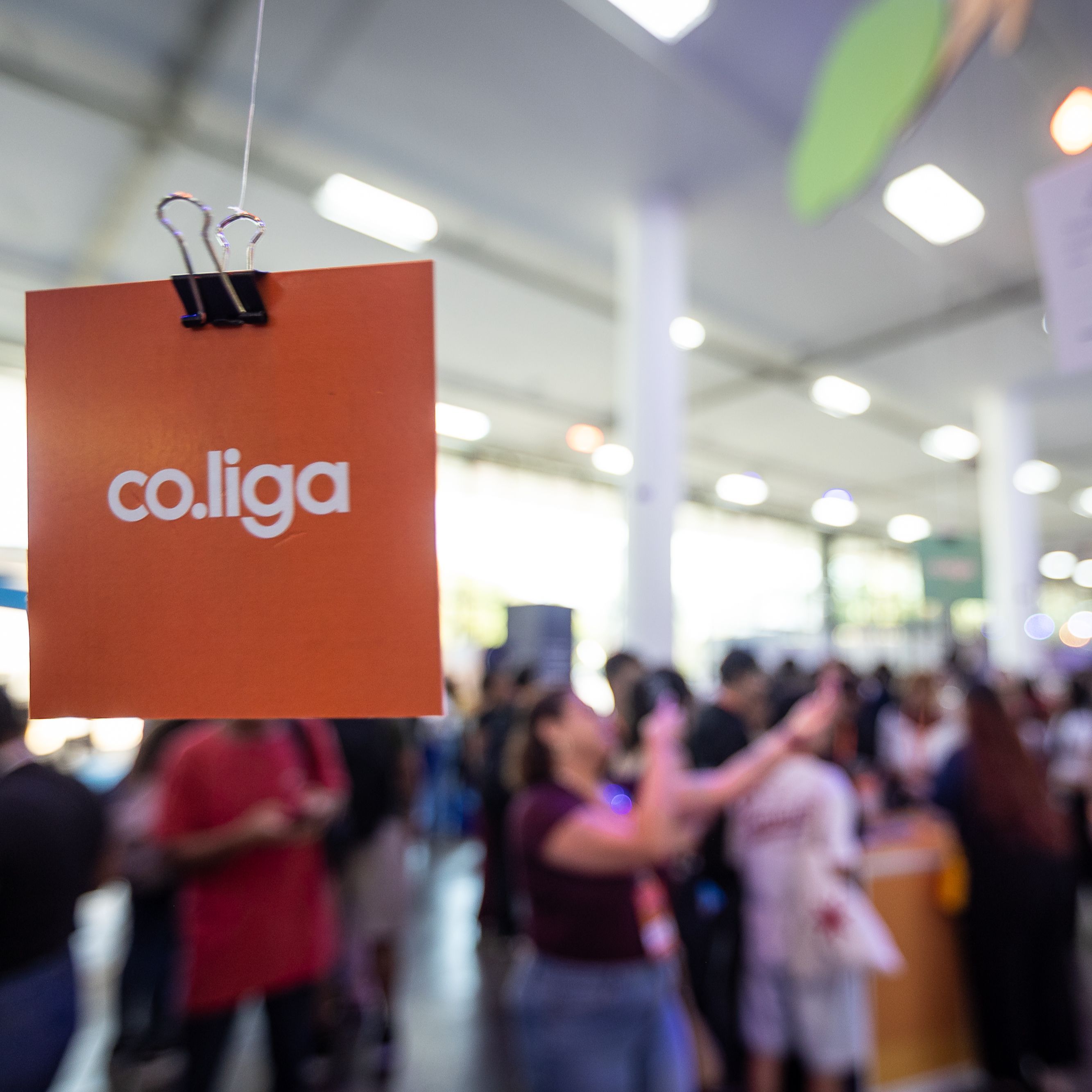Fotografia em primeiro plano de um cart&atilde;o quadrado cor de laranja suspenso por uma mola preta. No centro do cart&atilde;o est&aacute; impressa a palavra 